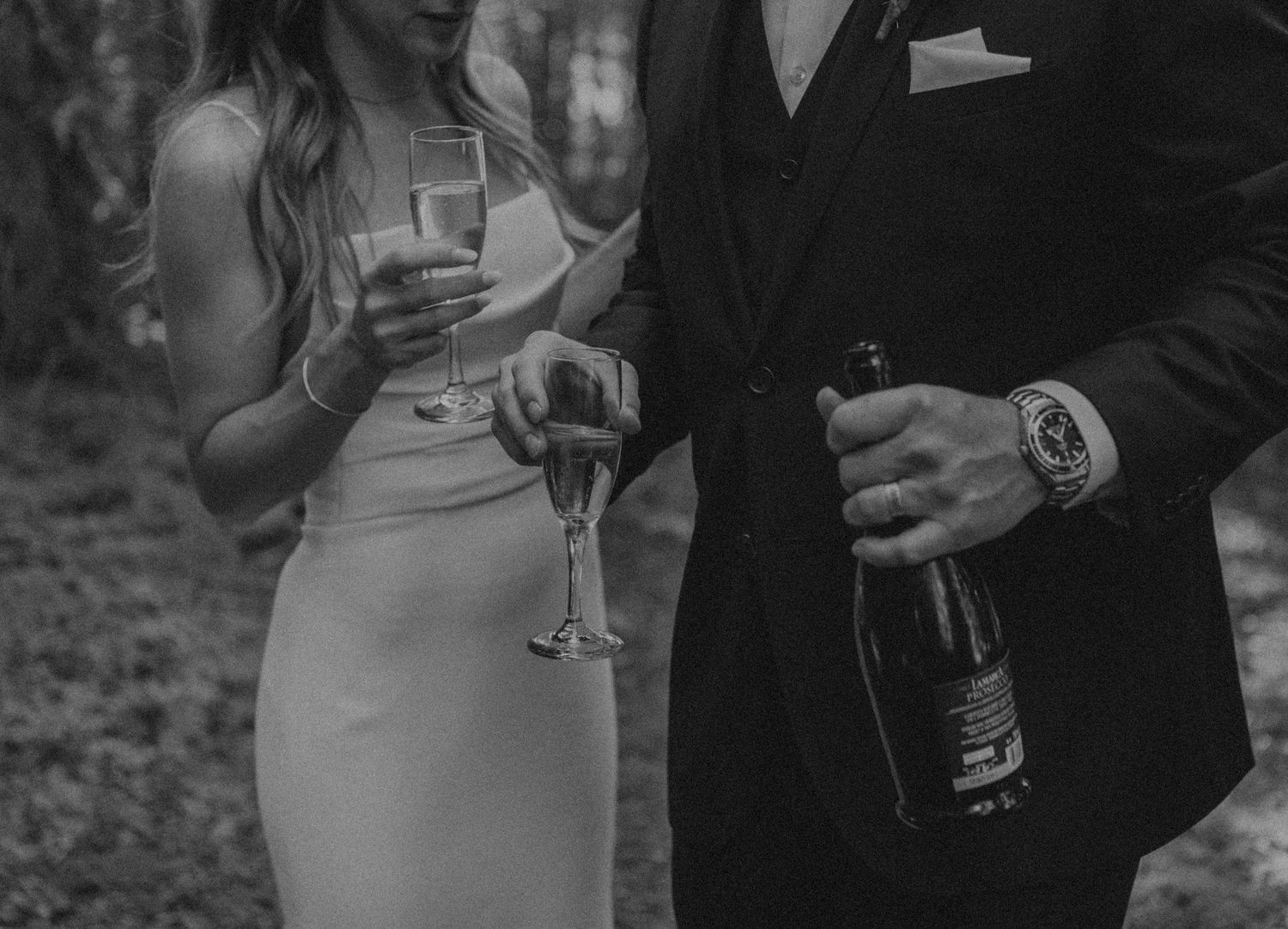 Black and white image of formal-dressed couple holding champagne glasses at an outdoor event.