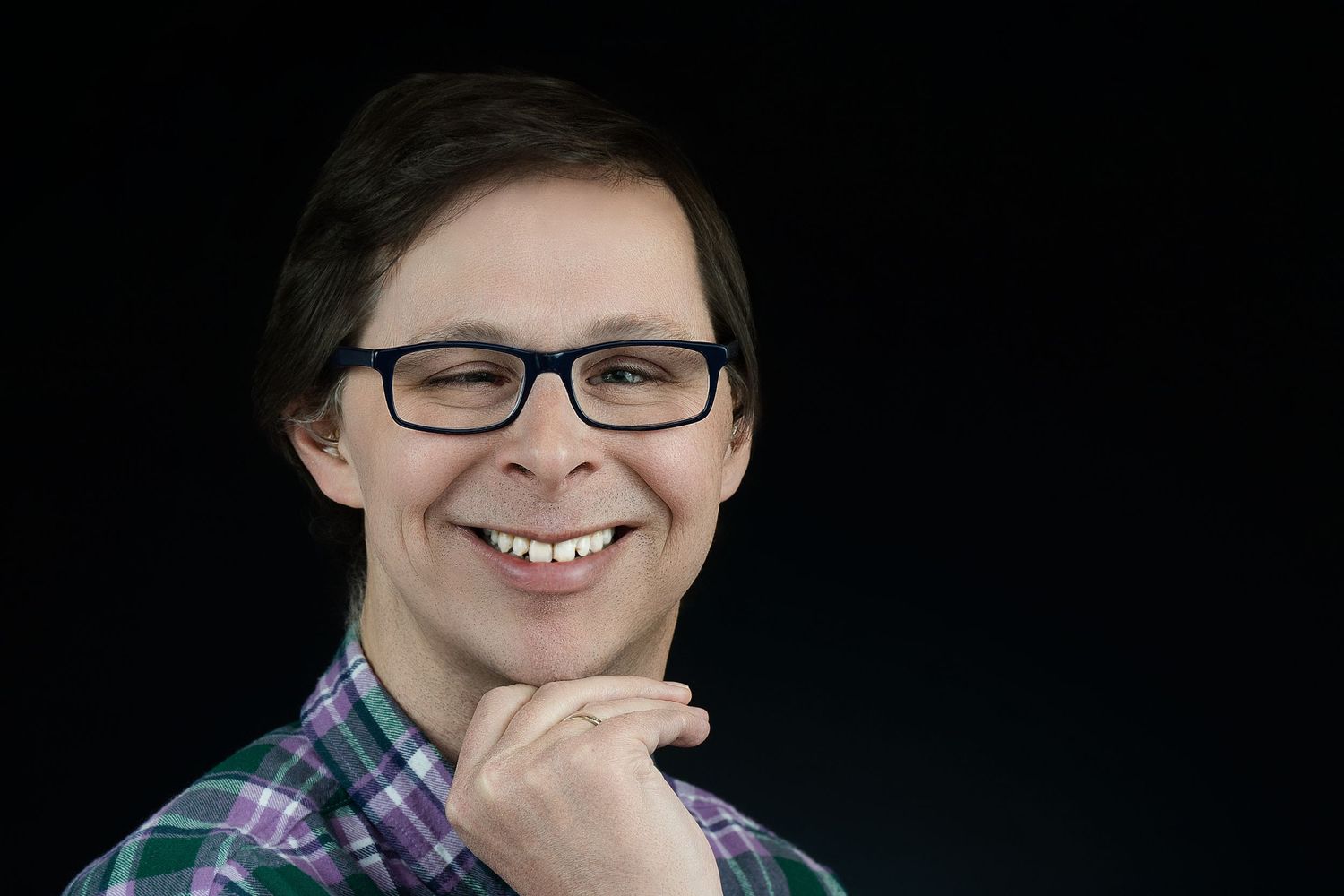 Portrait of person in plaid shirt and glasses with thoughtful expression against dark background.