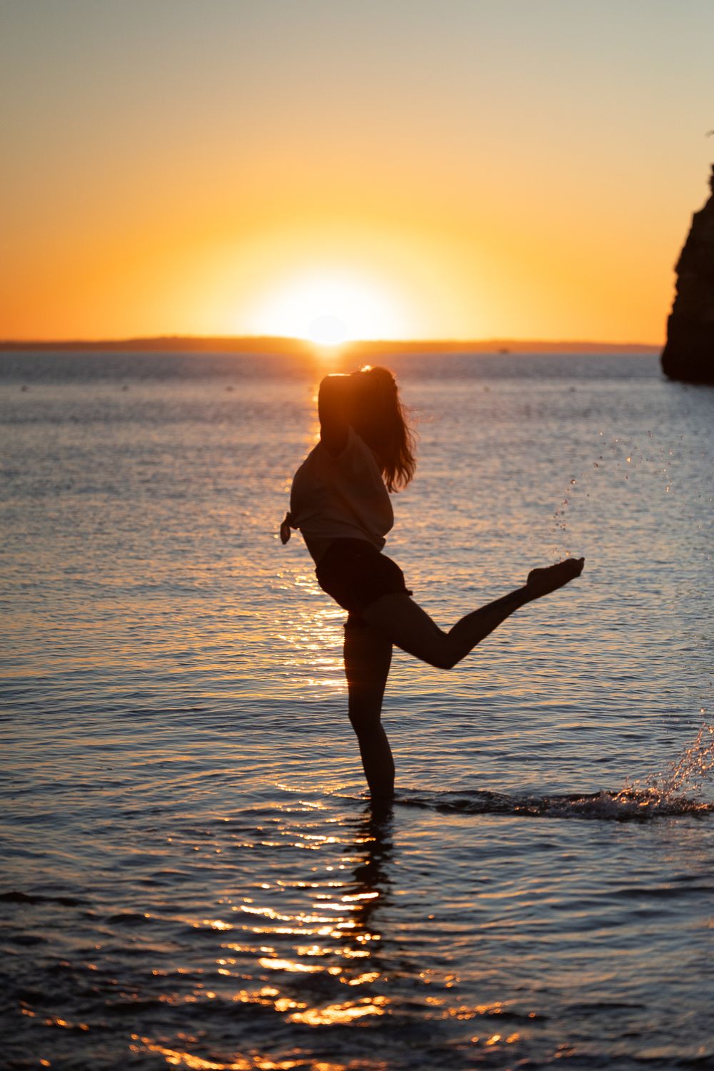 Silhouette of a woman kicking her leg in shallow ocean water during a vibrant golden sunset.