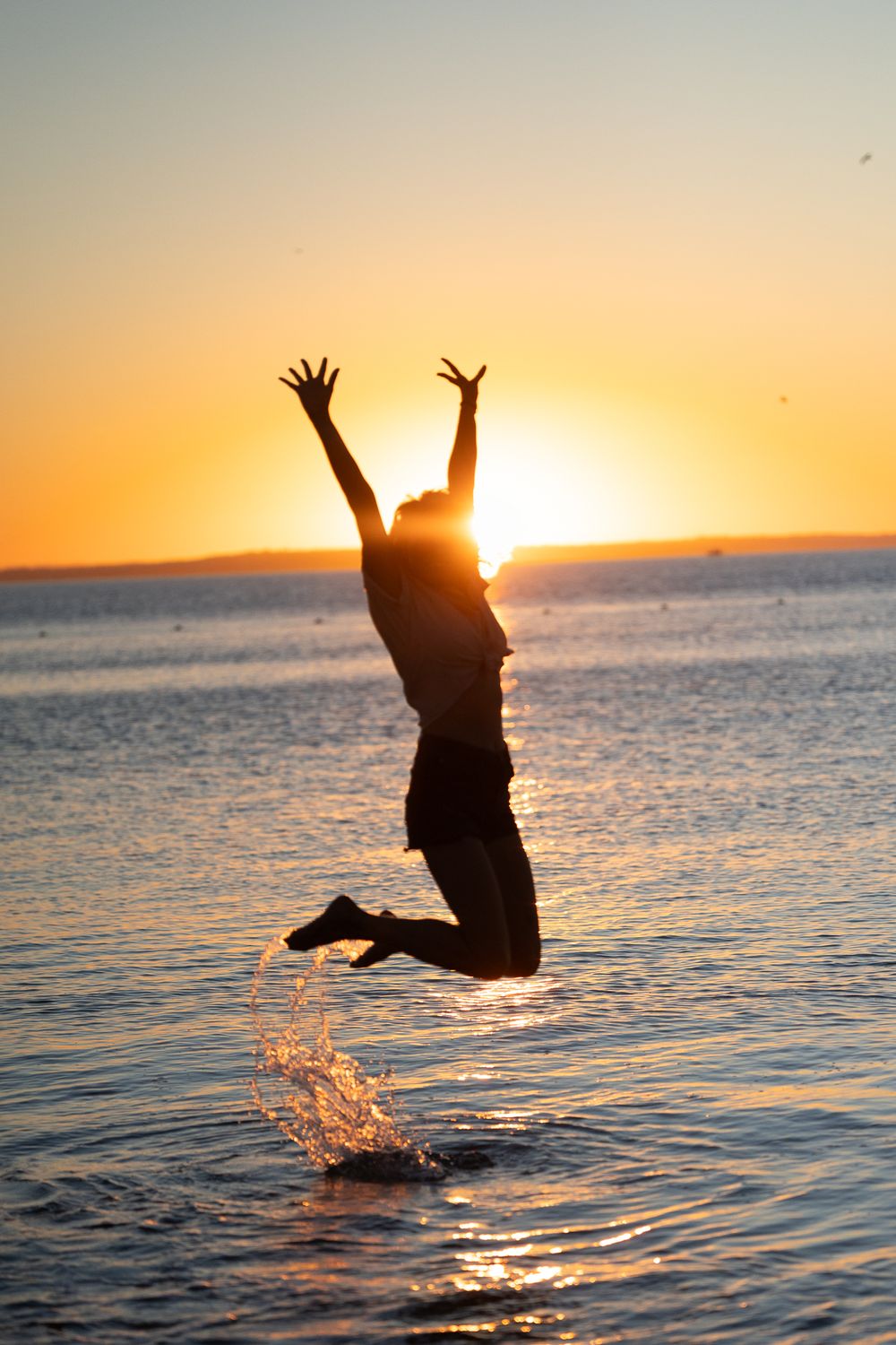Silhouette of a person jumping joyfully in ocean water during a vibrant golden sunset.
