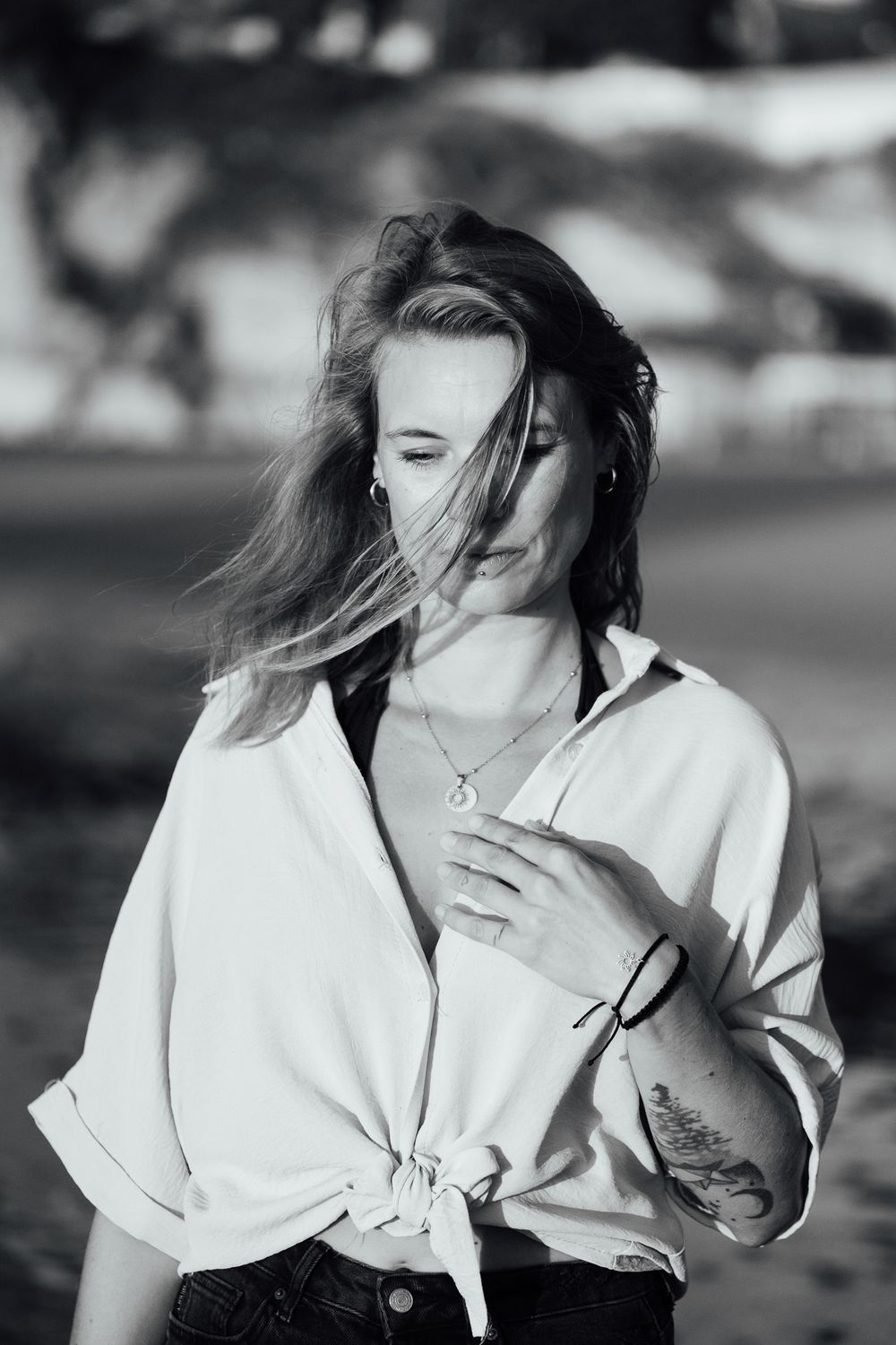 Black and white portrait of a young woman with wind-blown hair, wearing a tied white shirt and necklace outdoors.