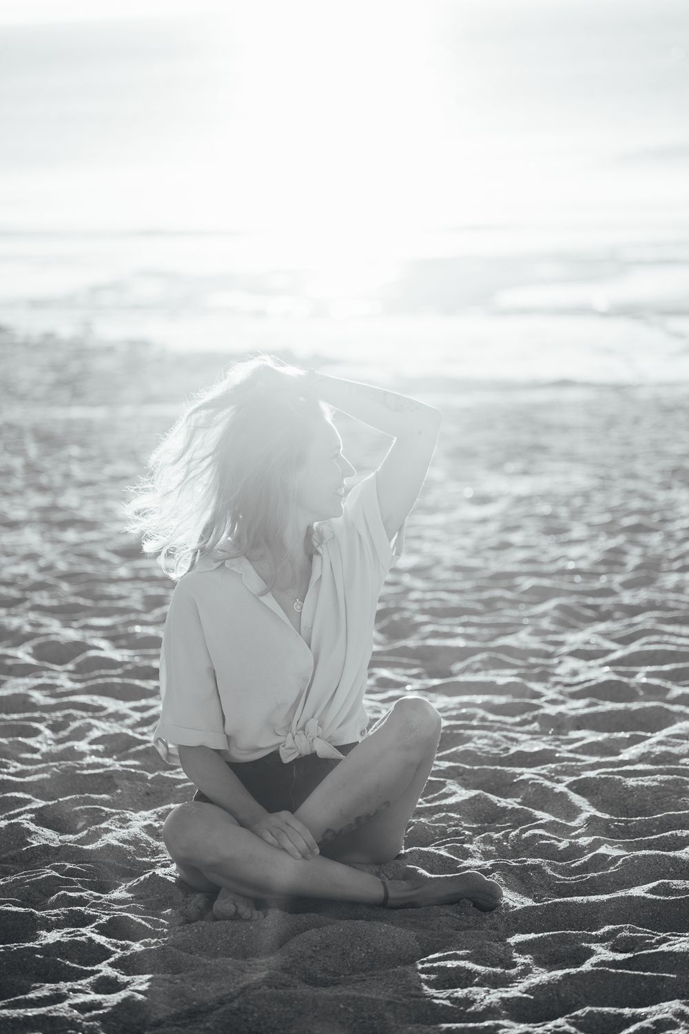Black and white photo of a woman sitting cross-legged on a sandy beach with wind-blown hair and bright sunlight.