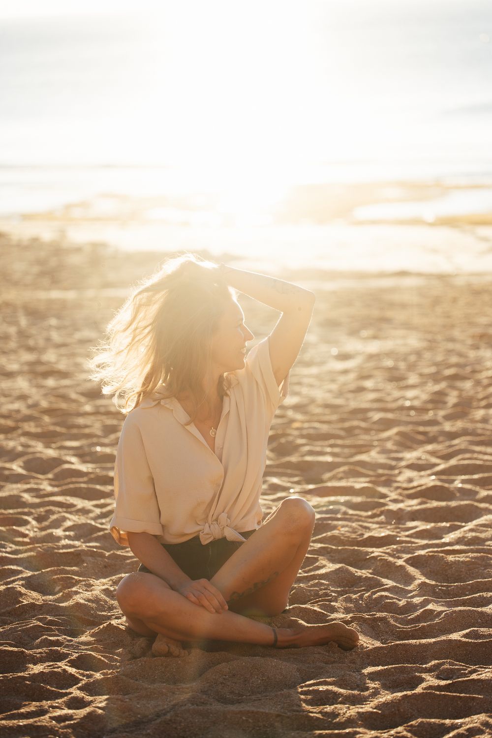 Woman sitting cross-legged on sandy beach at golden sunset, hand in windswept hair, wearing white shirt and shorts.