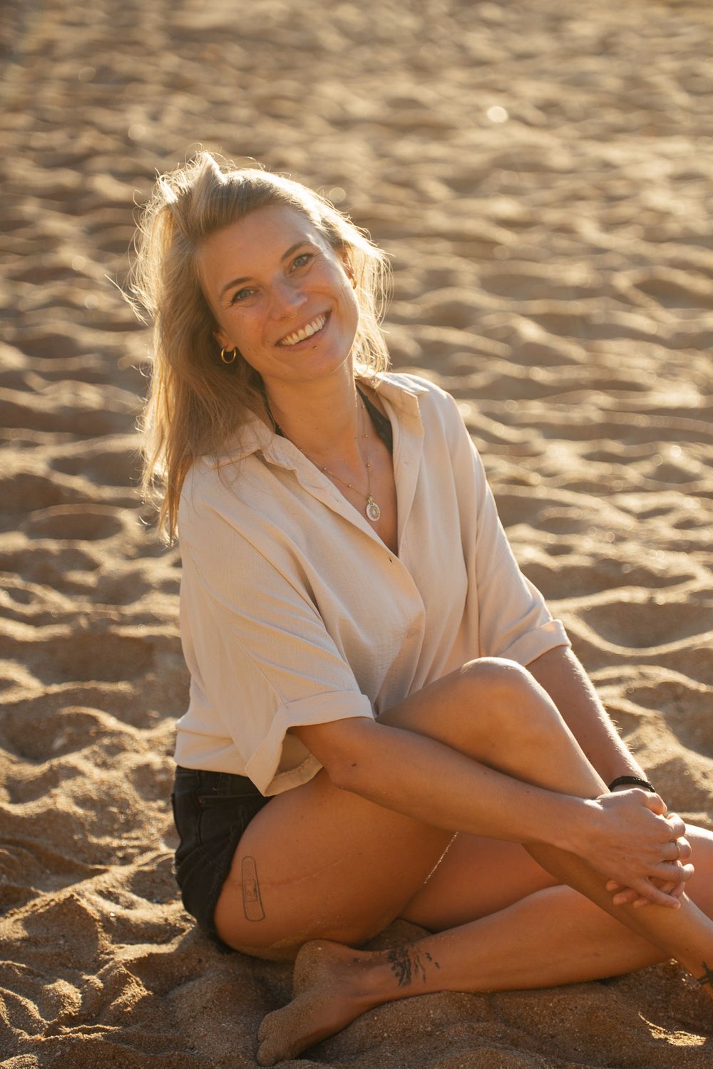 Smiling blonde woman sitting cross-legged on sandy beach in warm golden sunlight wearing white shirt.