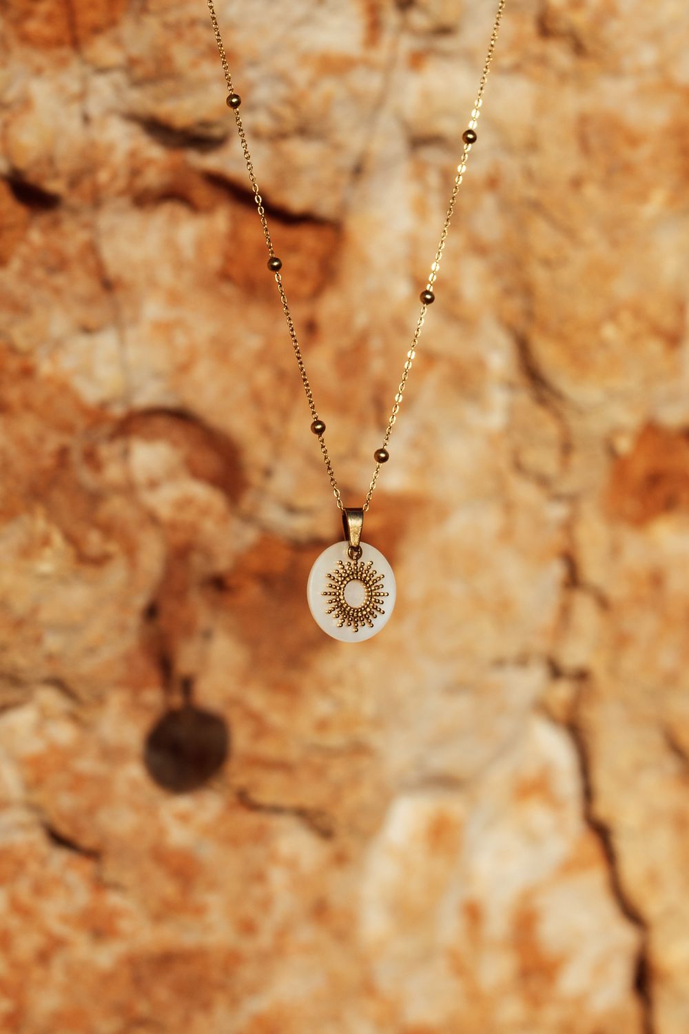 Gold chain necklace with a white circular pendant featuring a sun design, displayed against a rustic stone background.