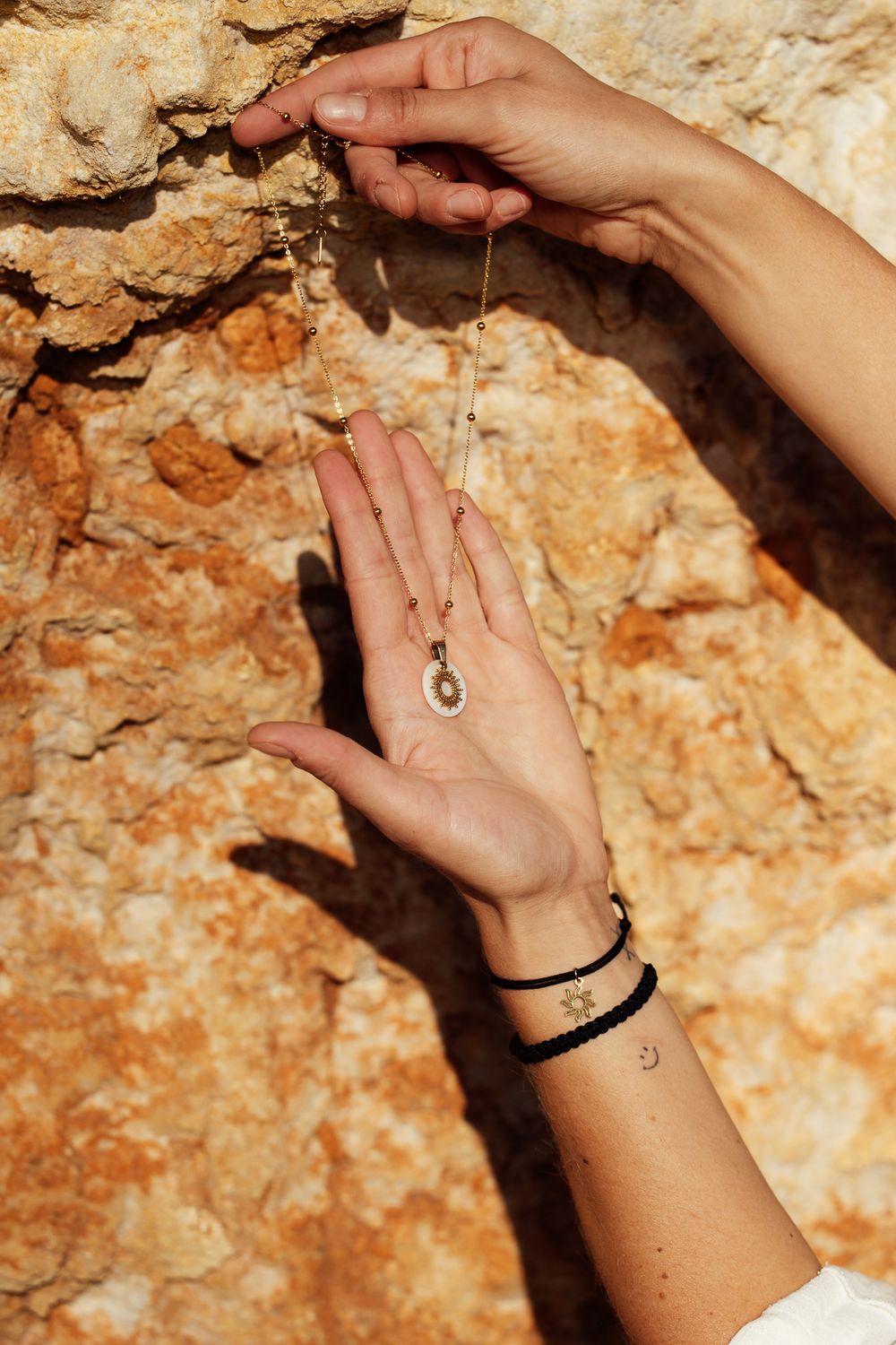 Hands displaying a delicate gold necklace with pendant, wearing black cord and star bracelets against a rocky stone wall.