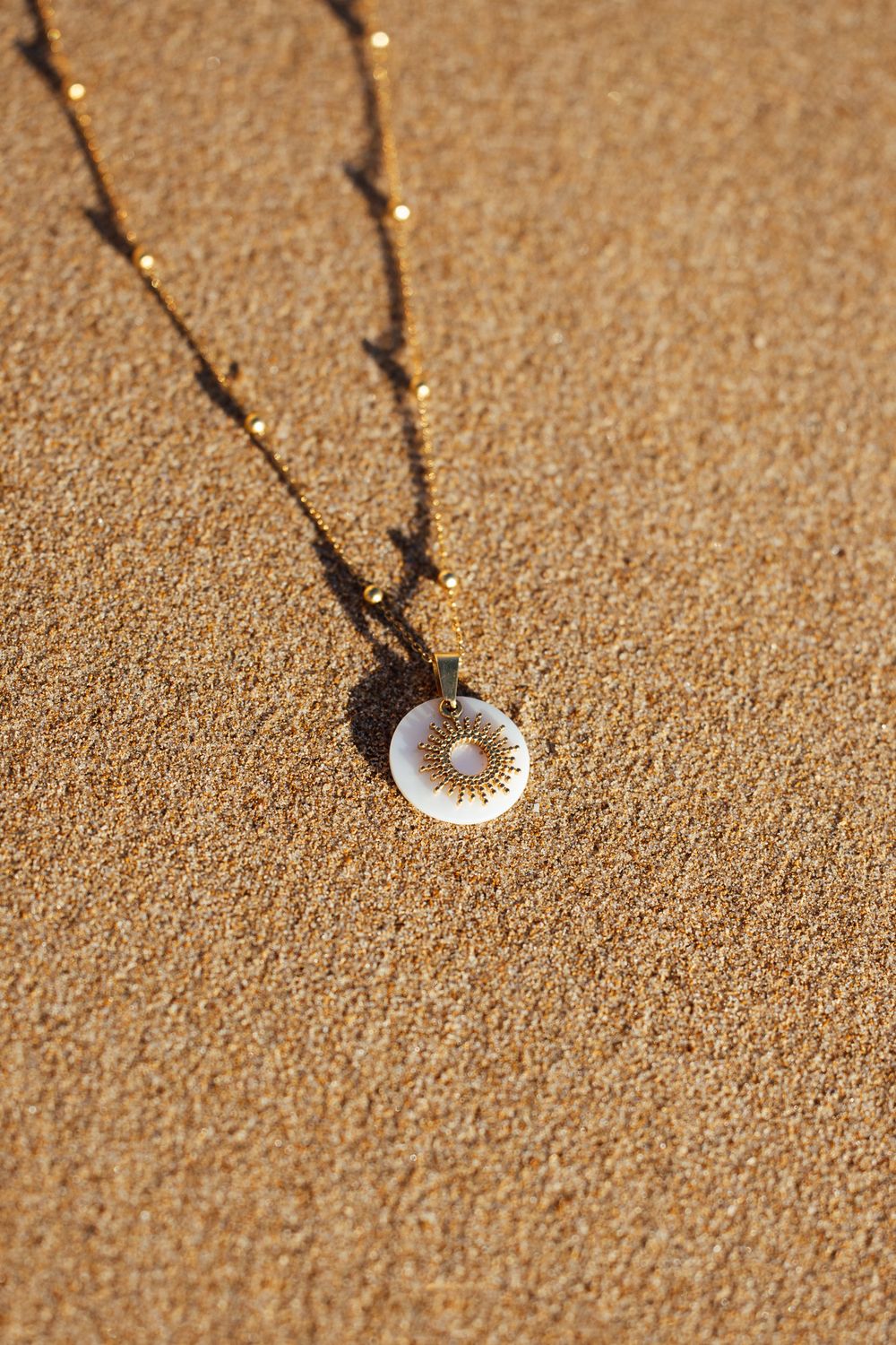 Gold chain necklace with a circular white shell sun pendant resting on golden sand beach.