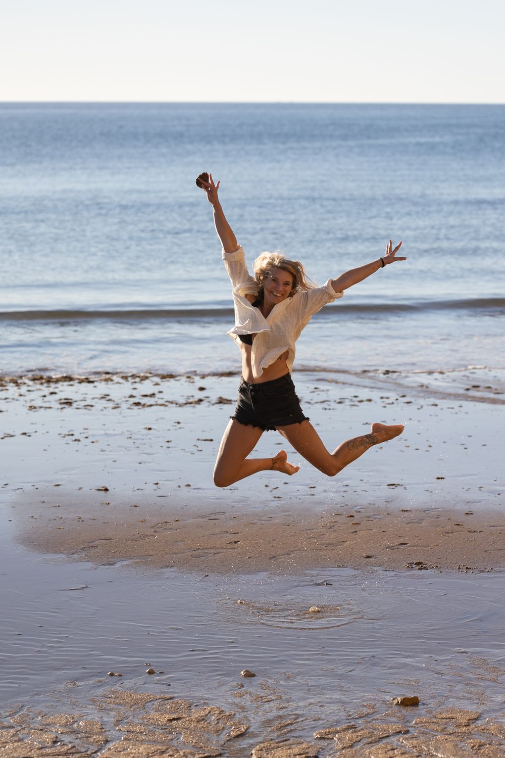 A joyful blonde woman jumps on a sandy beach with arms raised, wearing a white shirt and black shorts.