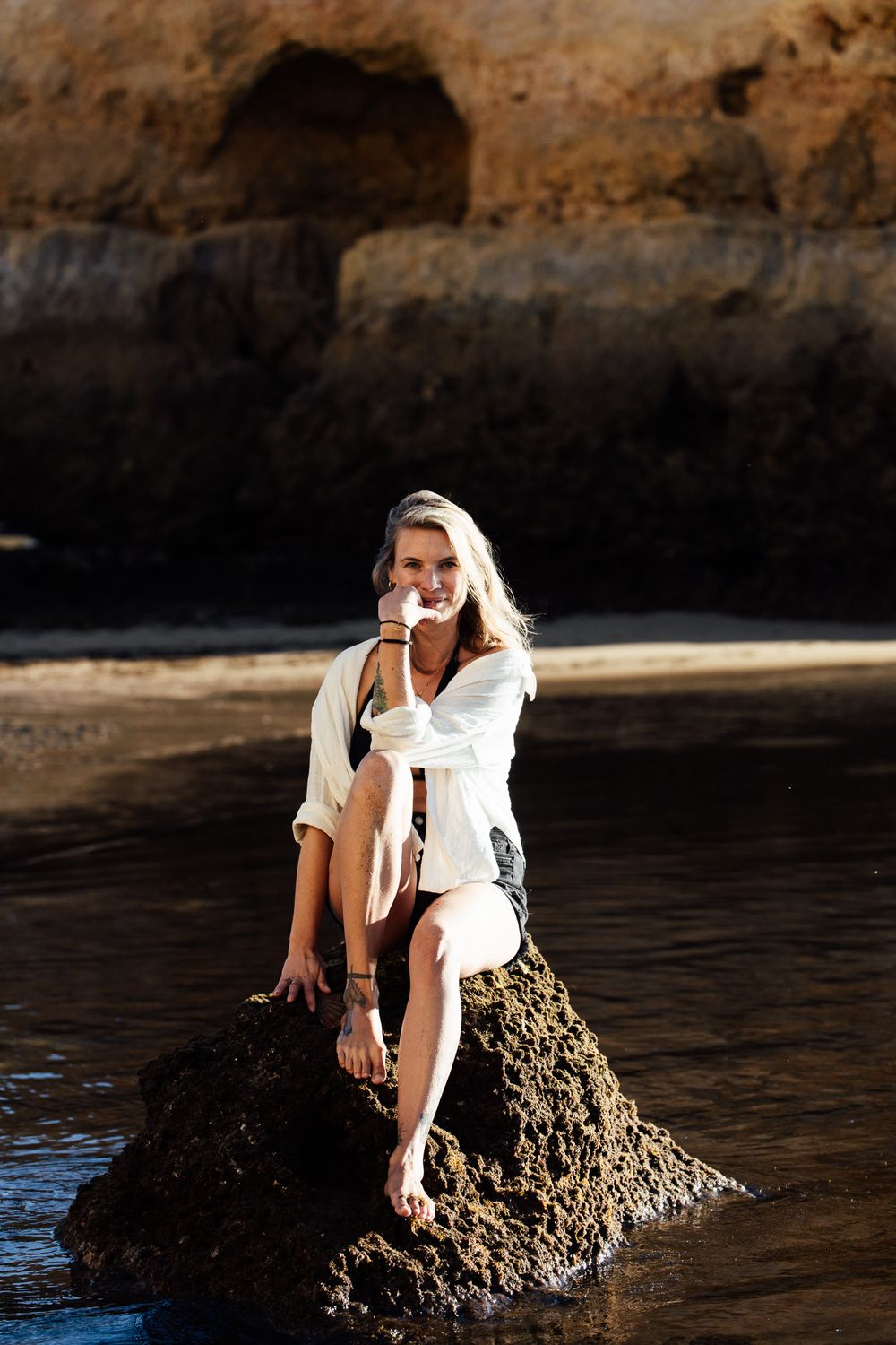 A smiling blonde woman sits on a rocky outcrop by the sea, wearing a white shirt and shorts, with cliffs behind her.