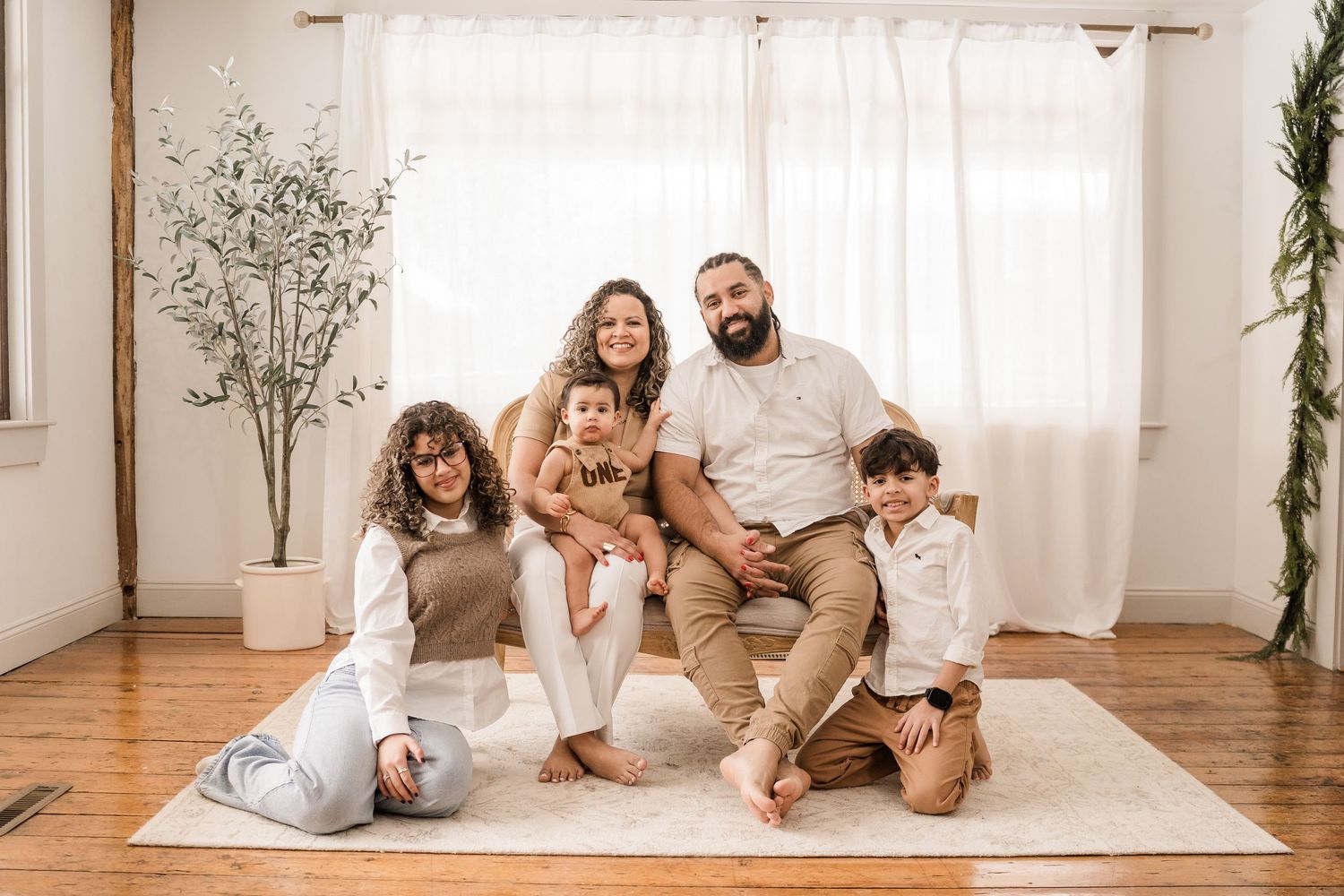 A family lounges together on a white blanket in a bright studio with wooden floors and potted plants.