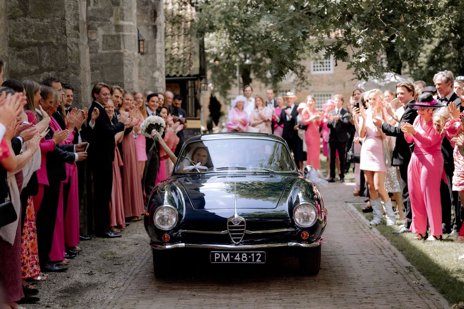 Wedding guests in pink attire line up to cheer for a classic black vintage car departing a stone building.