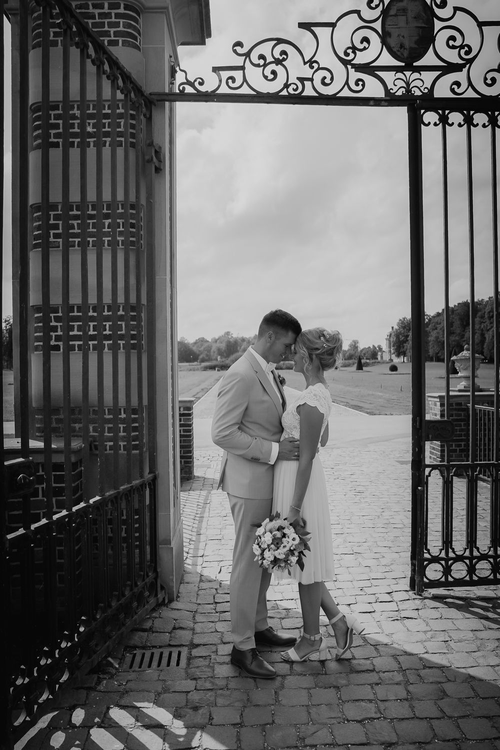 Newlywed couple sharing romantic moment by ornate iron gates at elegant venue entrance.