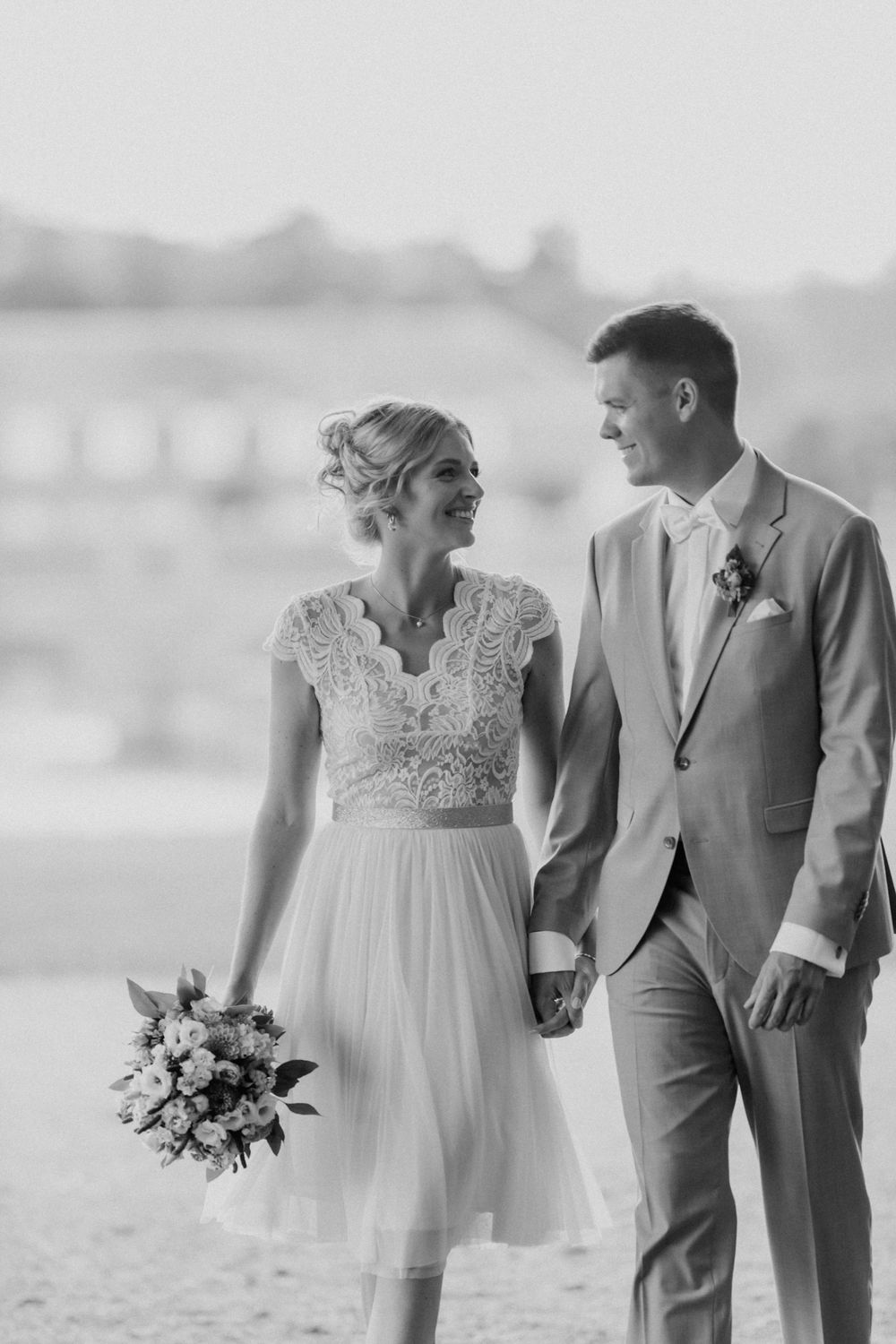 Black and white photo of newlyweds holding hands on beach with bride holding bouquet.