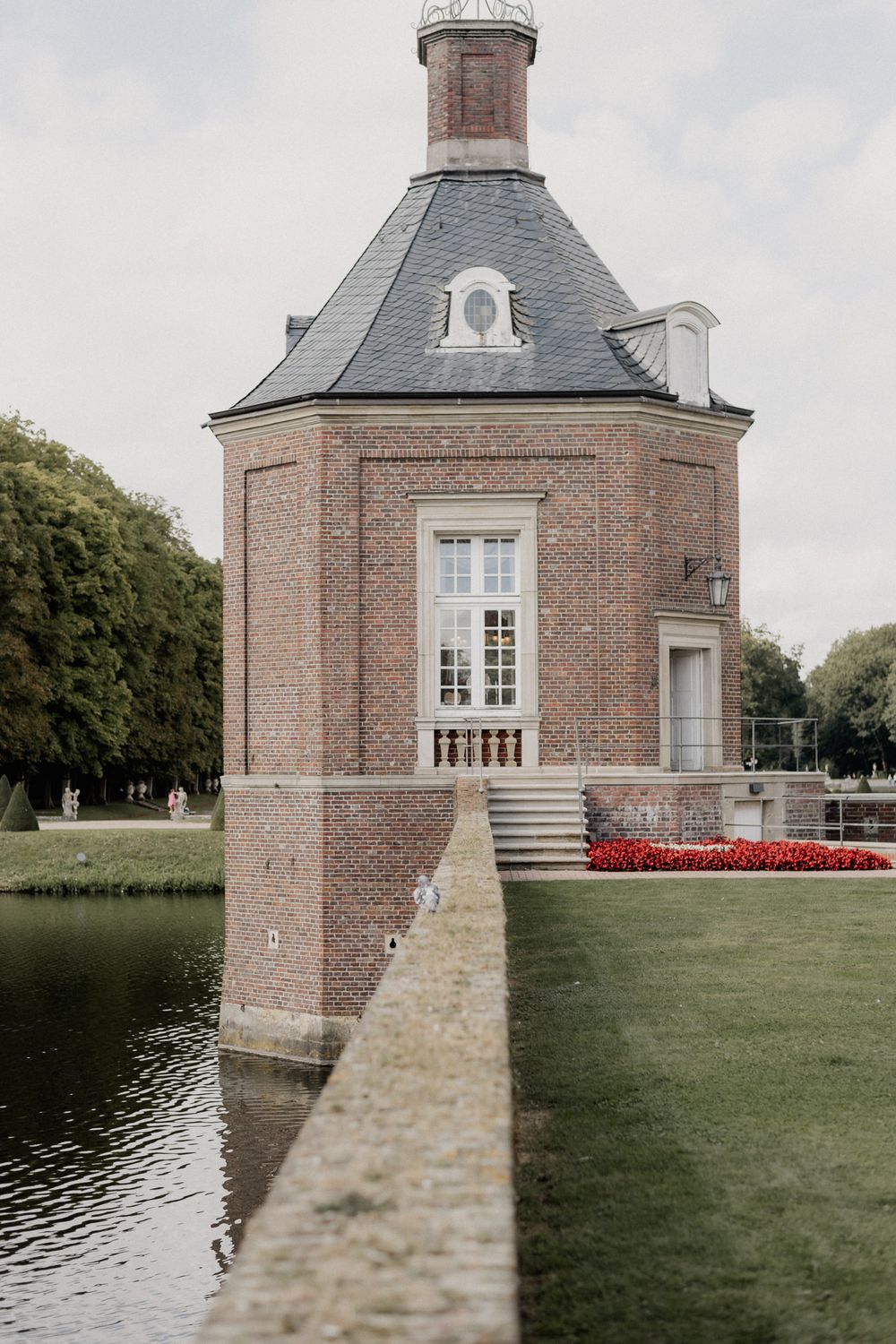 Historic brick pavilion with gray slate roof overlooking a pond and manicured garden.