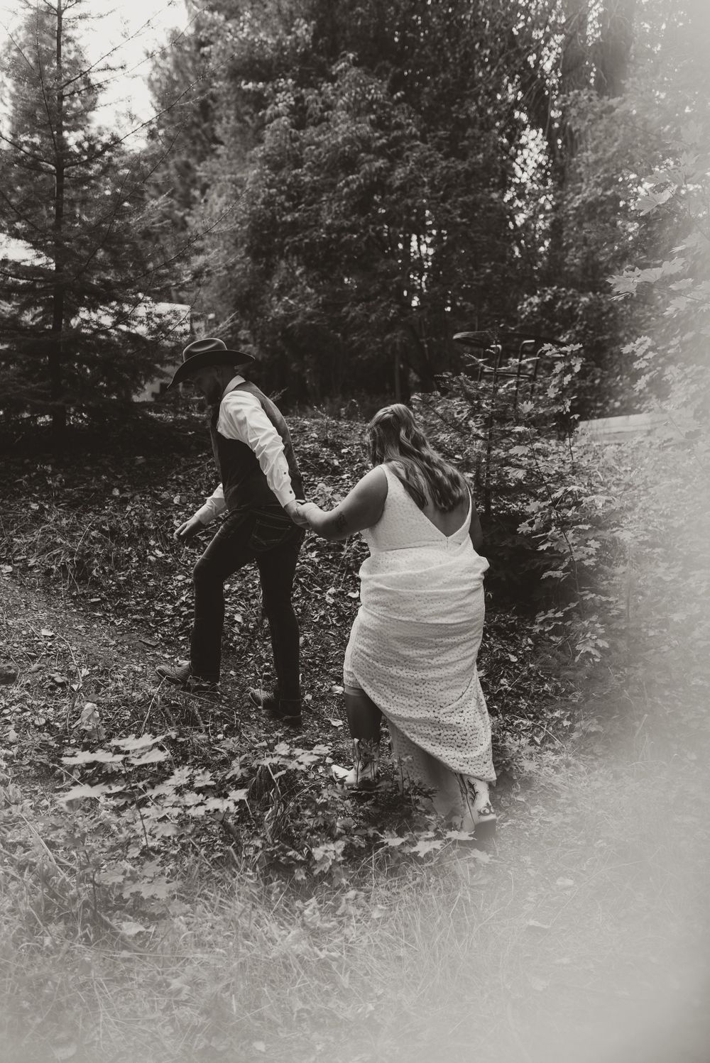 A black and white photo of a couple walking up a wooded hill in wedding attire.