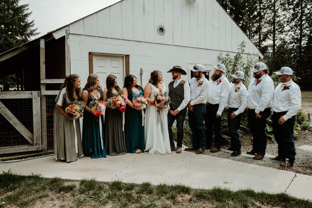 Wedding party lined up in rustic attire outside white barn building.