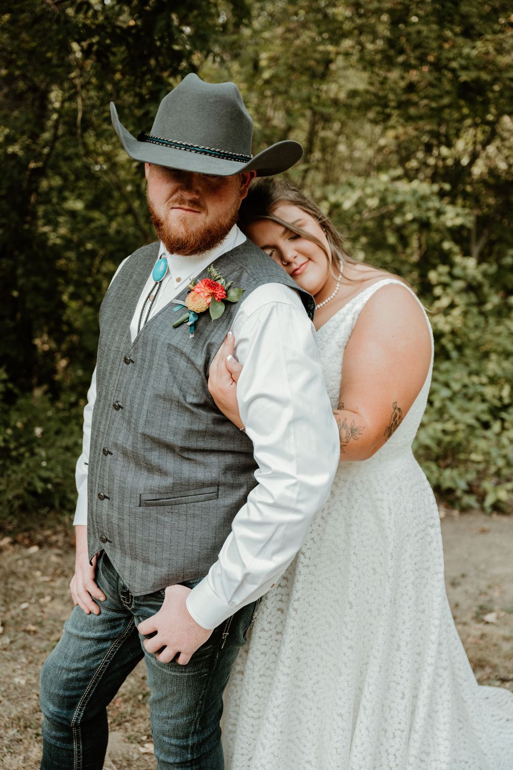 A couple embraces in western wedding attire with grey vest and white dress against a forest backdrop.