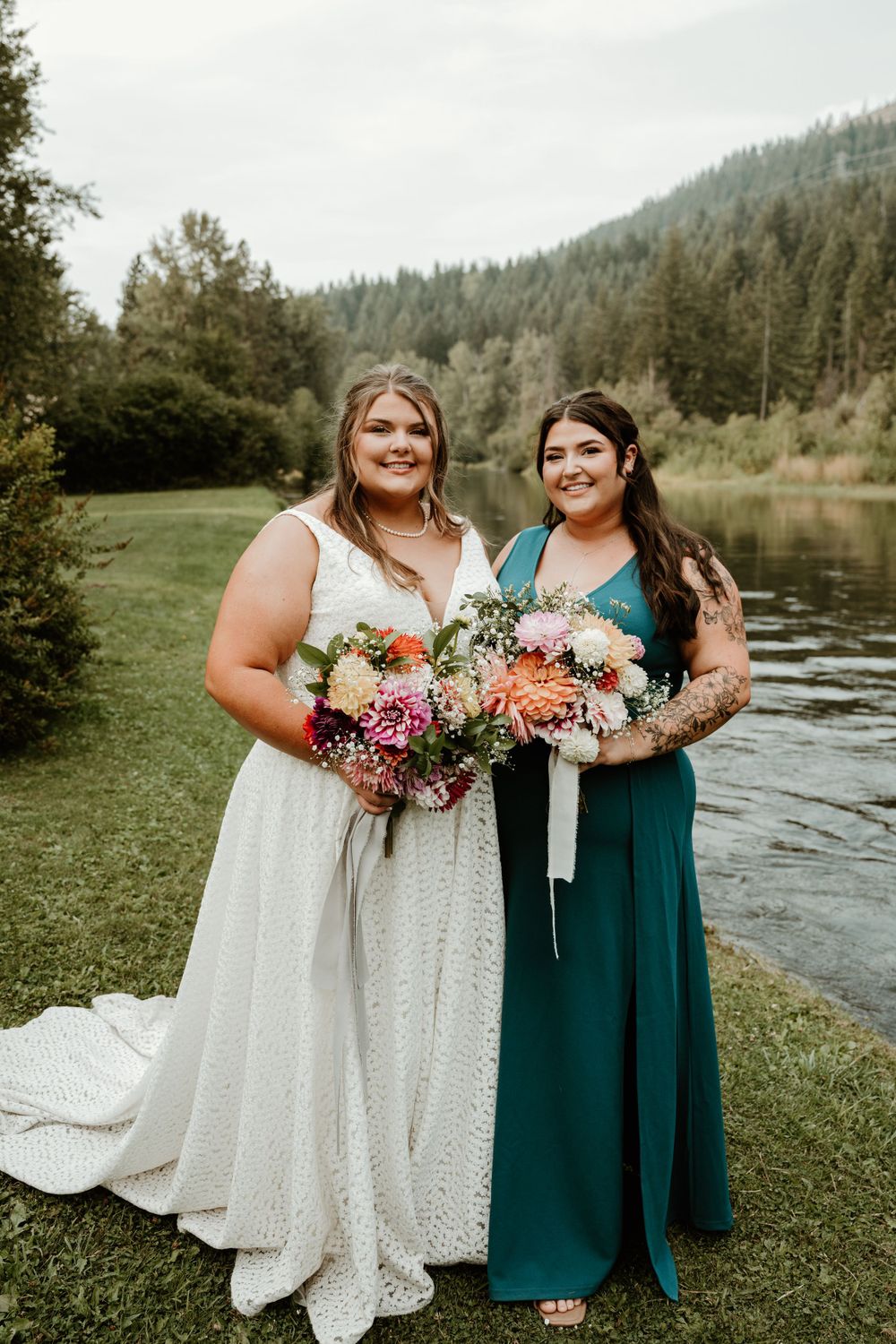 Two women in formal dresses hold colorful bouquets while standing by a scenic lake.