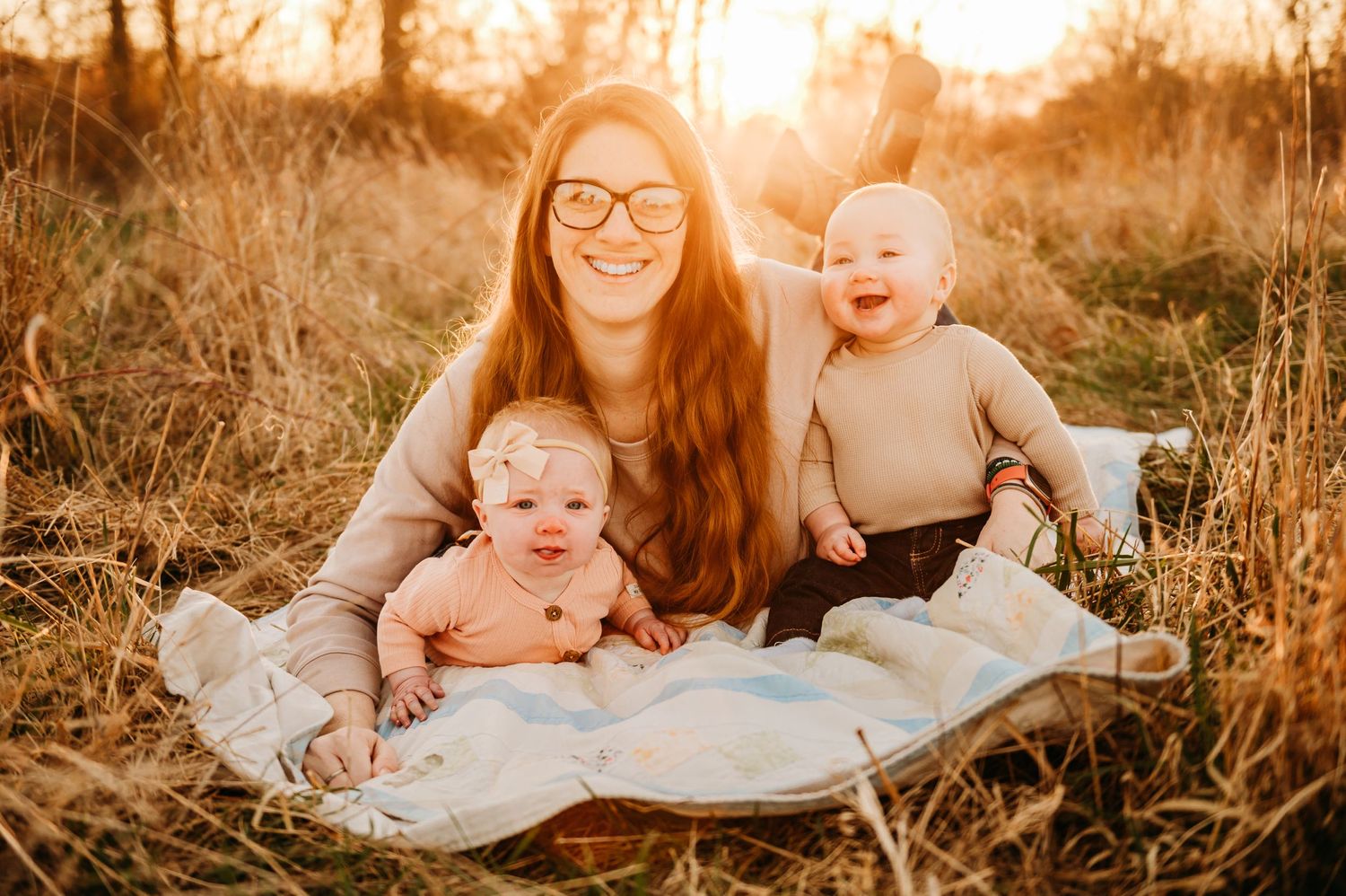 A golden sunset scene in a grassy field with three people sitting together on a white blanket during a fall afternoon.