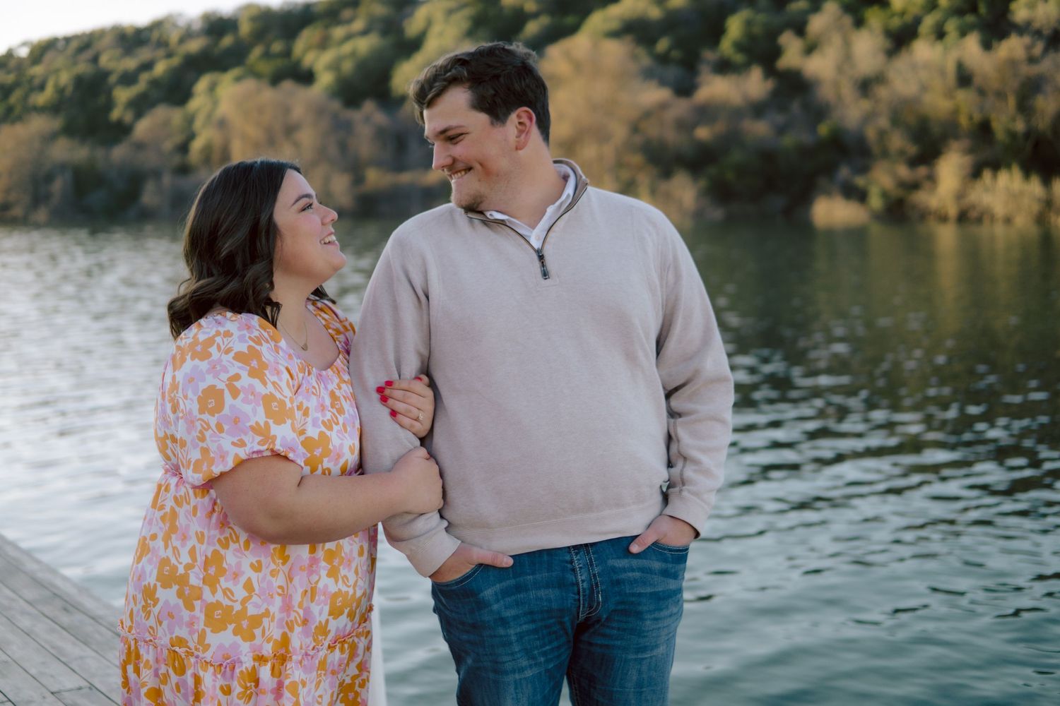 A couple stands lovingly by calm Lake Austin at sunset wearing a floral dress and beige sweater.