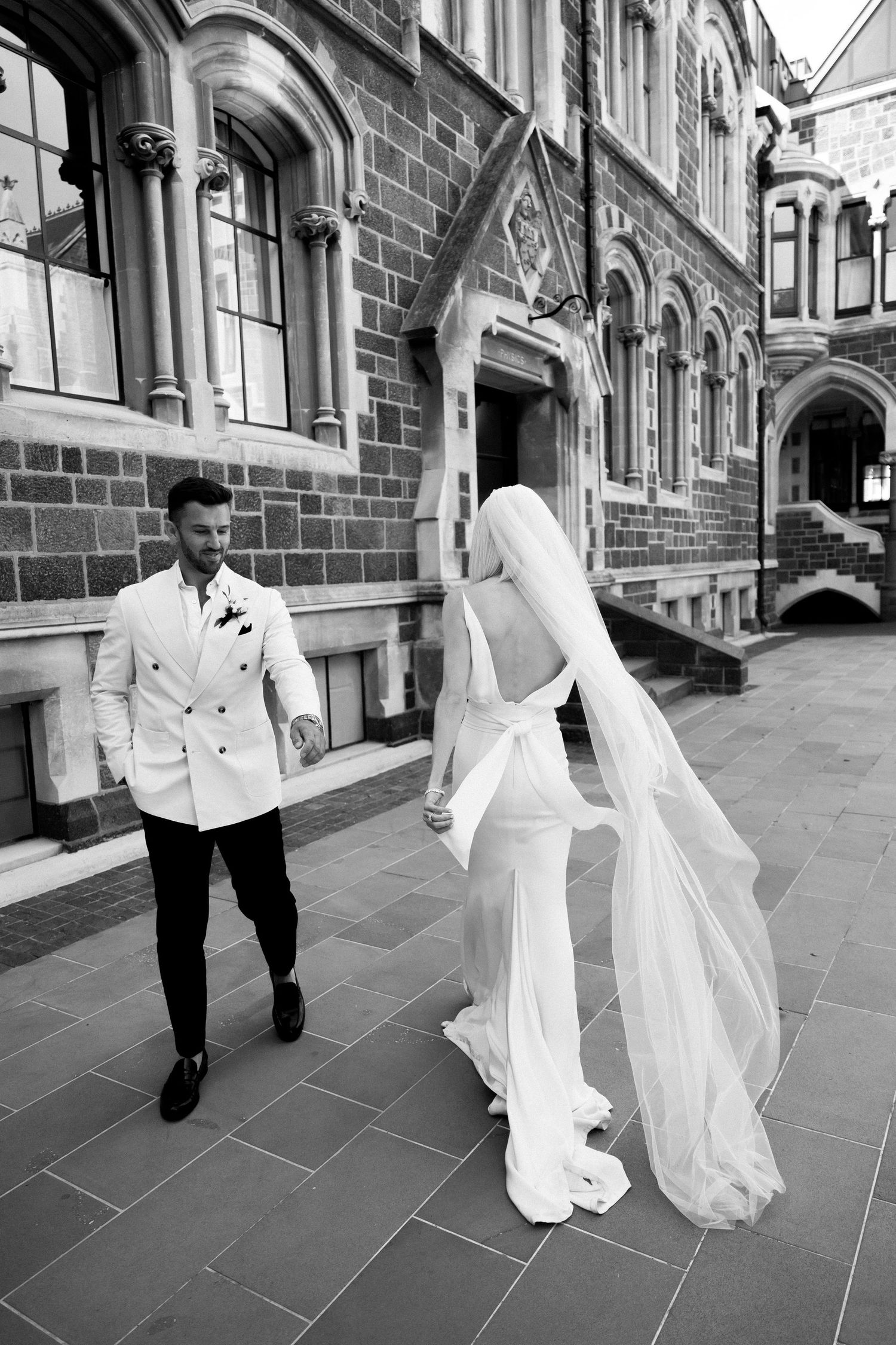 A couple in wedding attire stands outside a Gothic stone building with arched architecture and beautiful stonework.