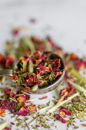 Colorful dried herbs and flower petals scattered with measuring spoons on a white surface.