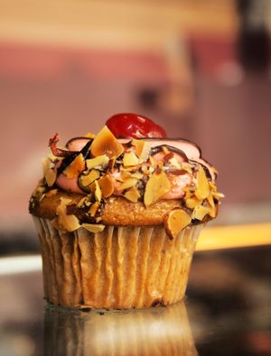 Gourmet cupcake topped with nuts, chocolate drizzle and cherry on reflective surface.