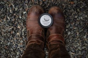 Brown leather boots on gravel with a round product tin placed between them.