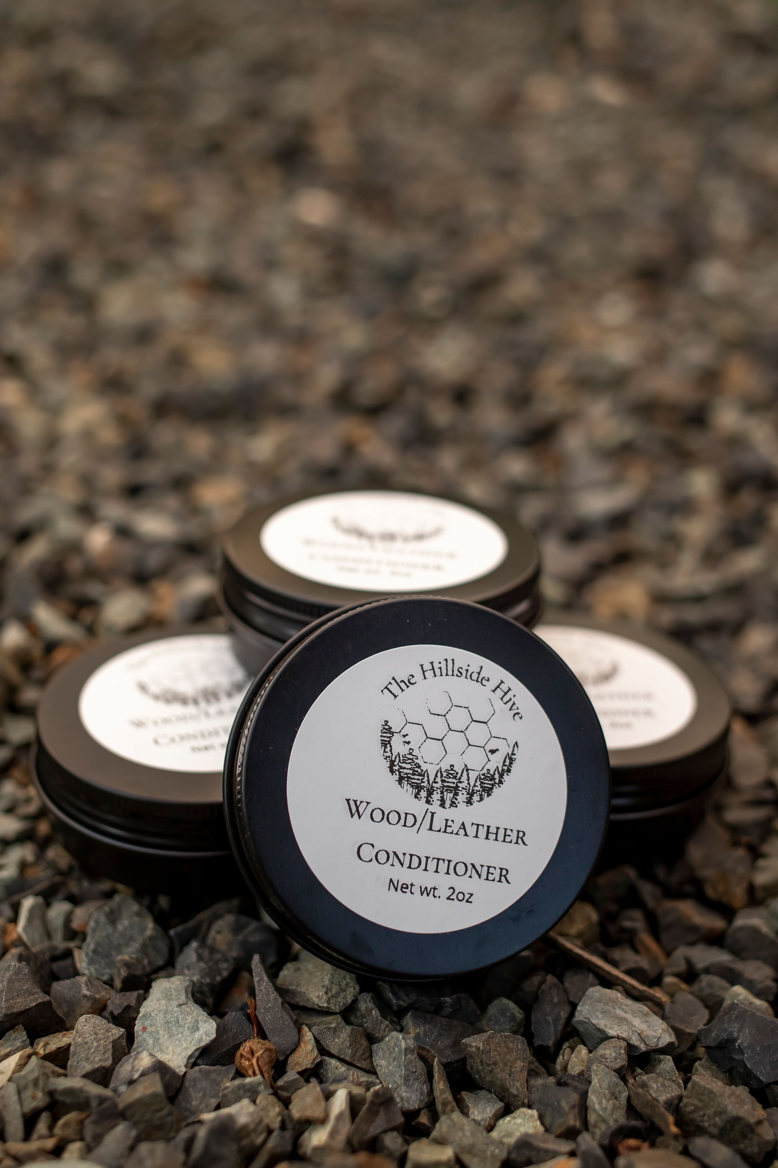Wood and leather conditioner tins with mountain logo displayed on gravel surface.