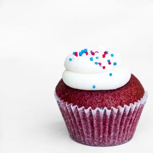Red velvet cupcake topped with white frosting and red and blue sprinkles on white background.