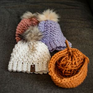 Cozy winter knit beanies with fur pom poms in white, lavender, coral and orange colors displayed on dark fabric.