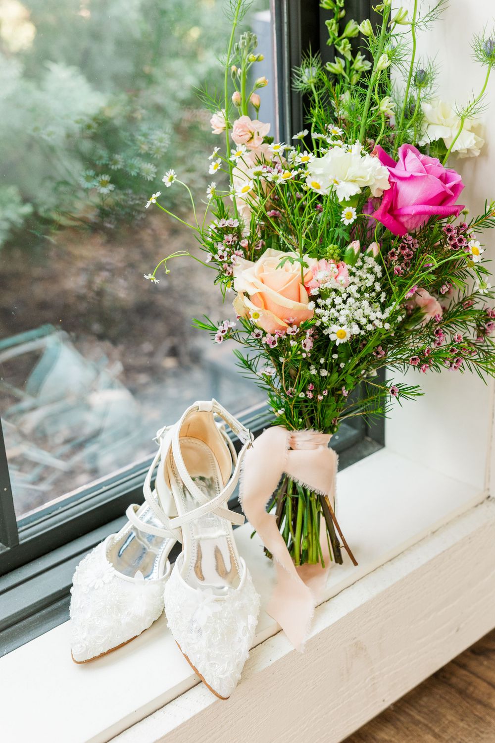 Elegant wedding bouquet with pink and peach roses next to white lace bridal shoes on a windowsill.