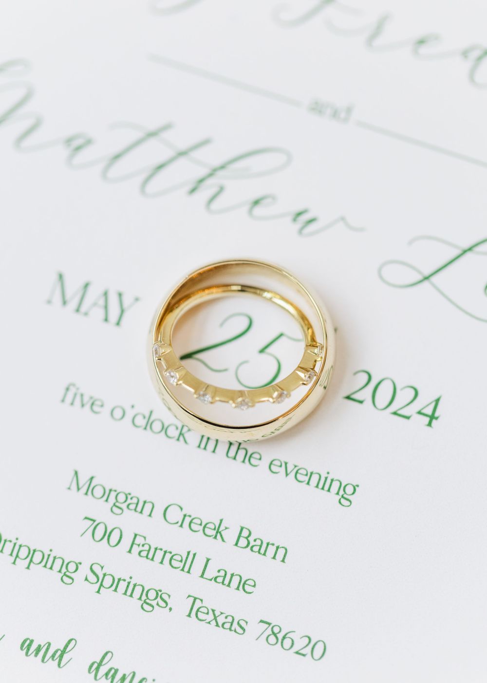 Gold wedding rings rest on an elegant green and white wedding invitation showing details for Morgan Creek Barn.