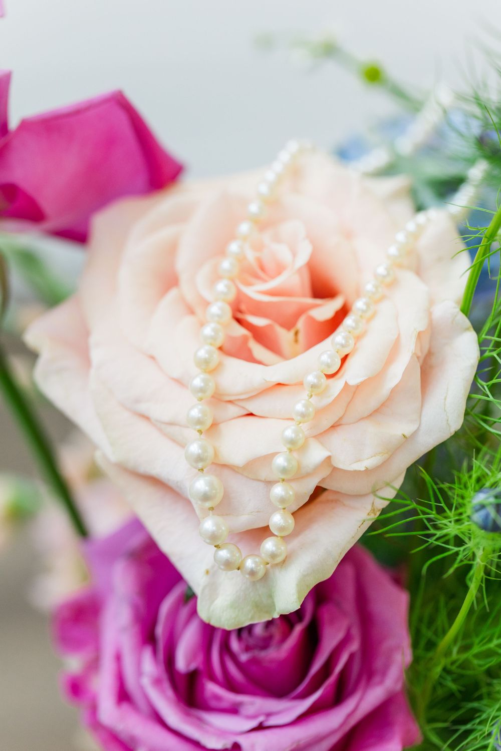 A delicate peach rose adorned with a strand of pearl beads against a background of pink flowers and greenery.