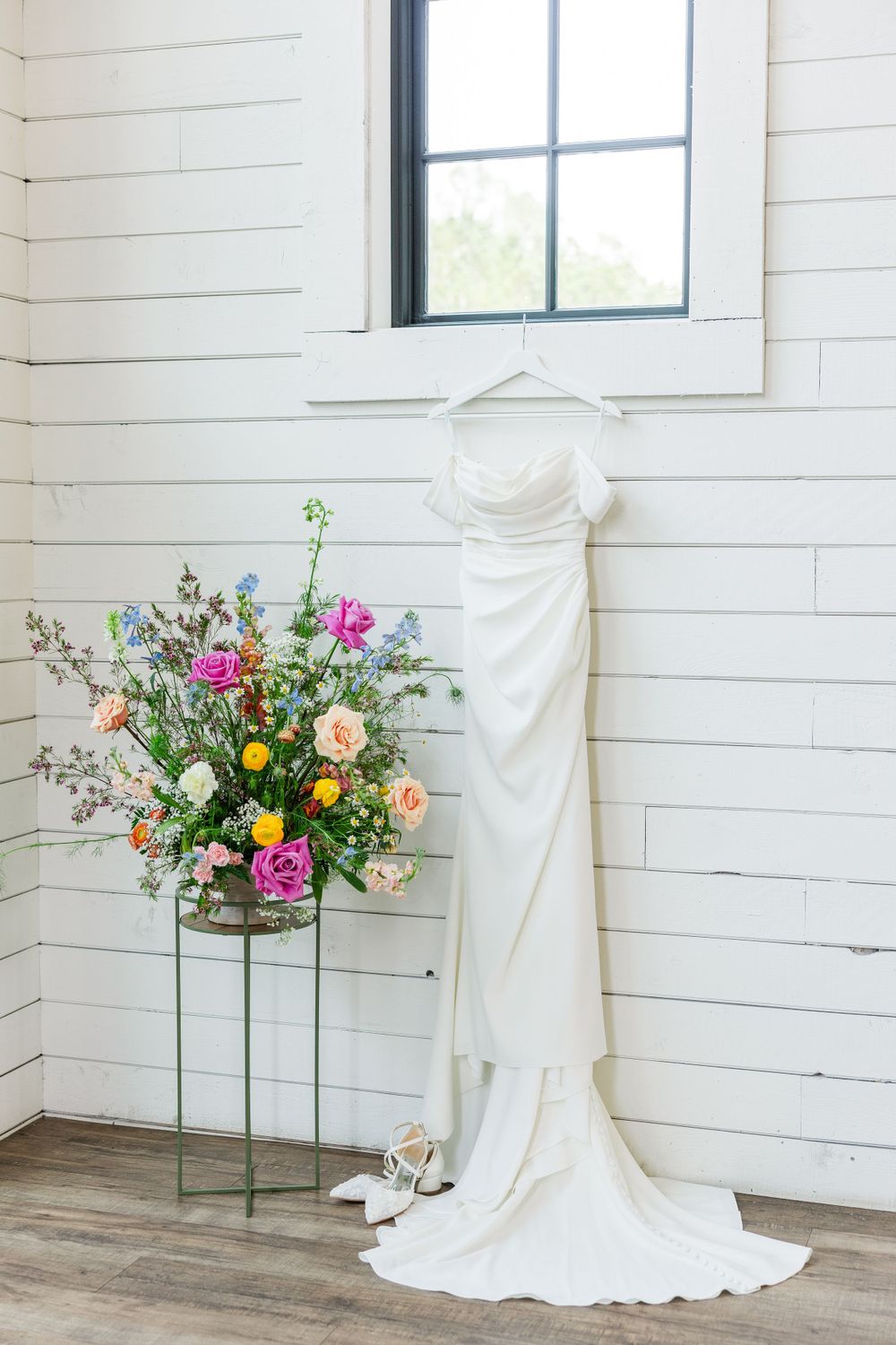 Wedding dress hanging on white shiplap wall next to colorful floral arrangement.