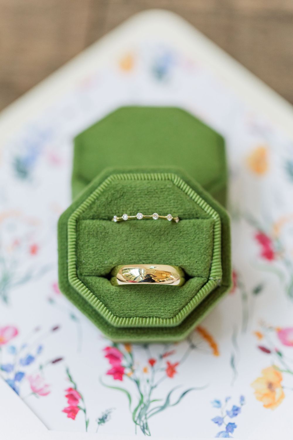 Gold wedding ring displayed in green velvet octagonal box on floral patterned paper.