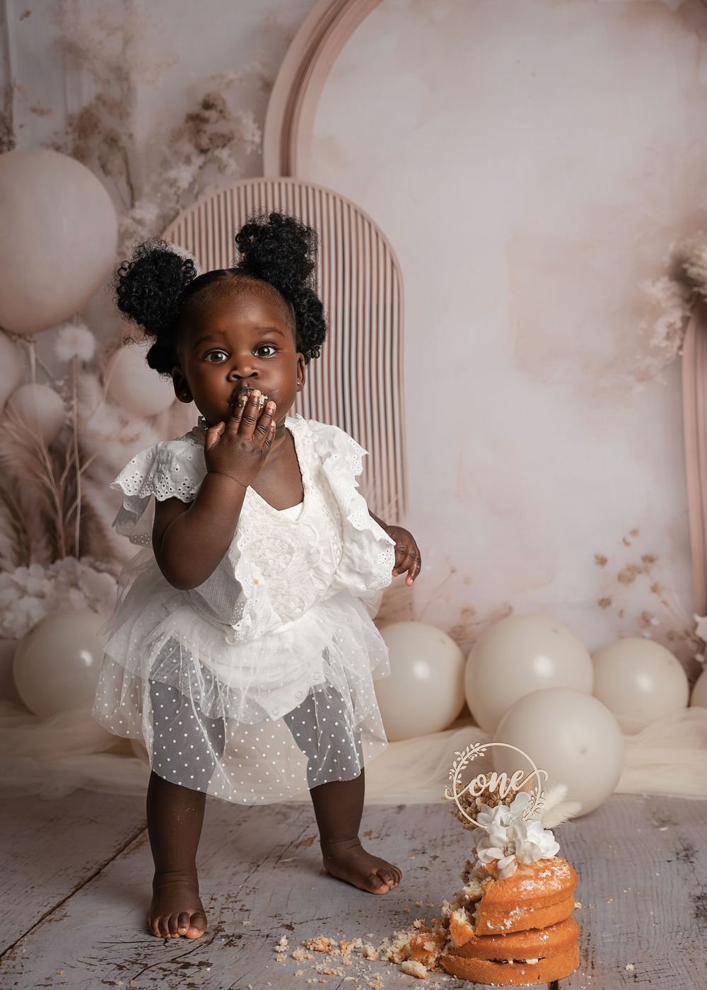 A toddler in a white lace dress sits beside a smashed cake and balloons in a dreamy pink studio setting.