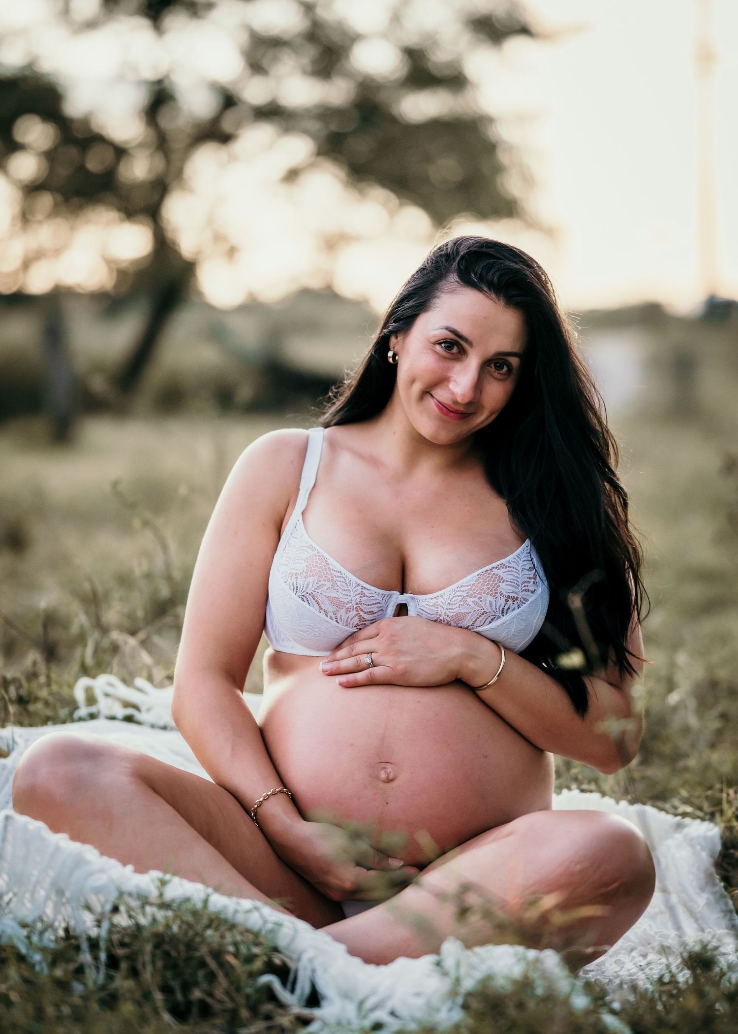 Natural maternity portrait of woman in white lace bra smiling at sunset in grassy field.