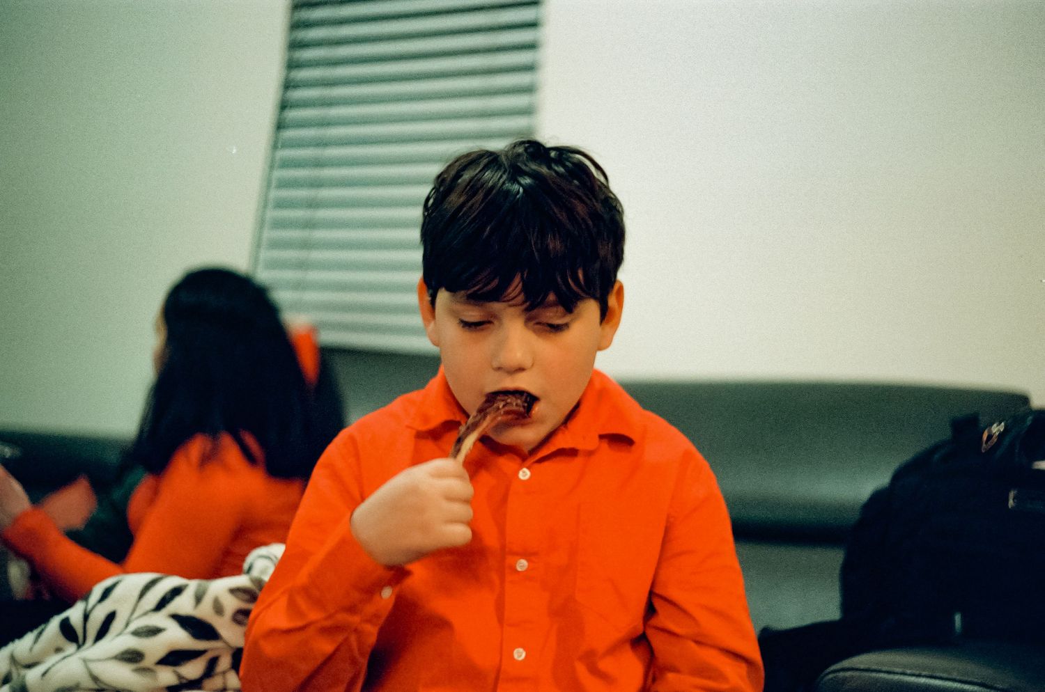 Young person in bright orange shirt eating or tasting something indoors.
