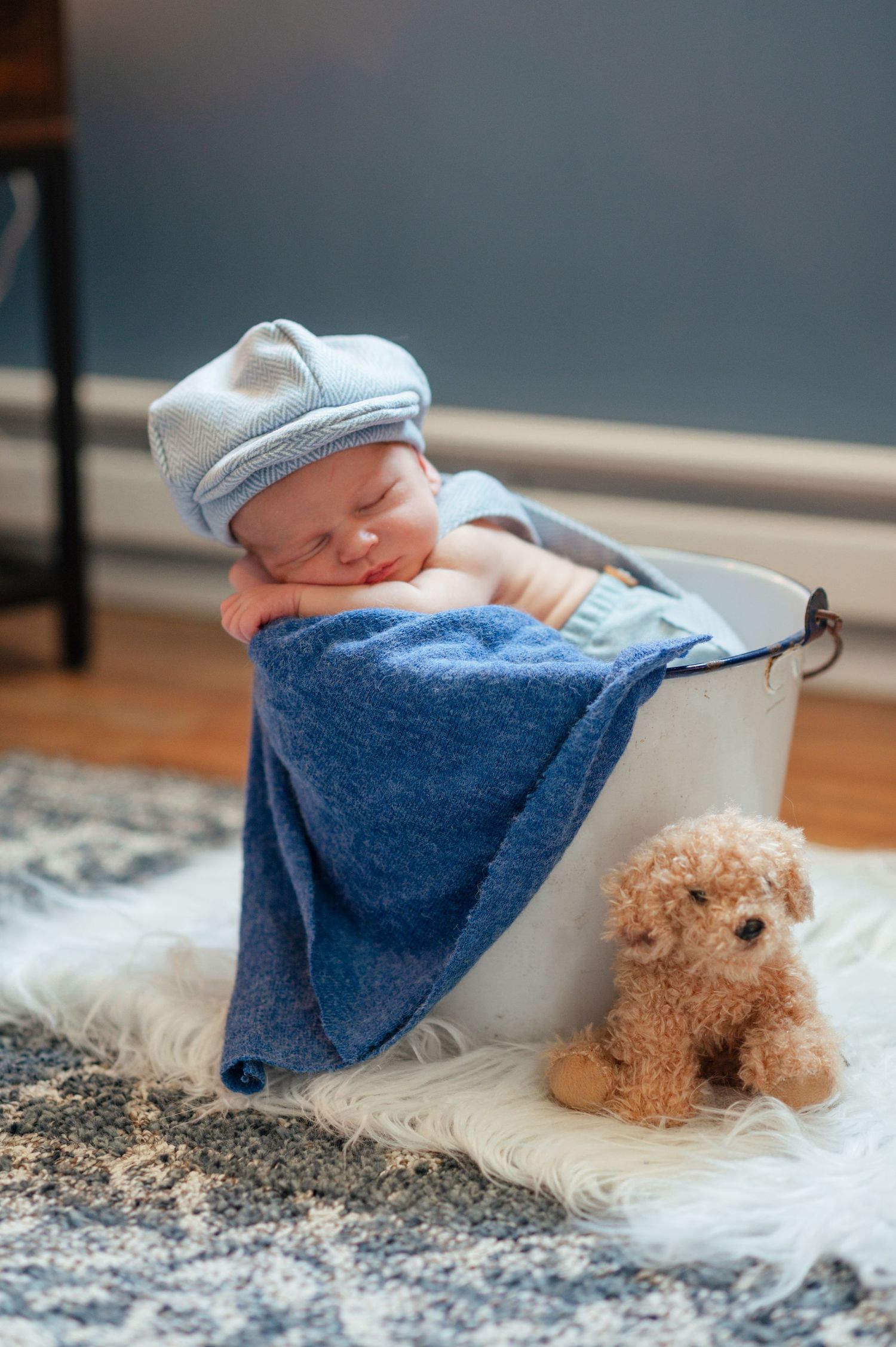 Newborn baby sleeping in white bucket with blue blanket and plush teddy bear on carpeted floor.
