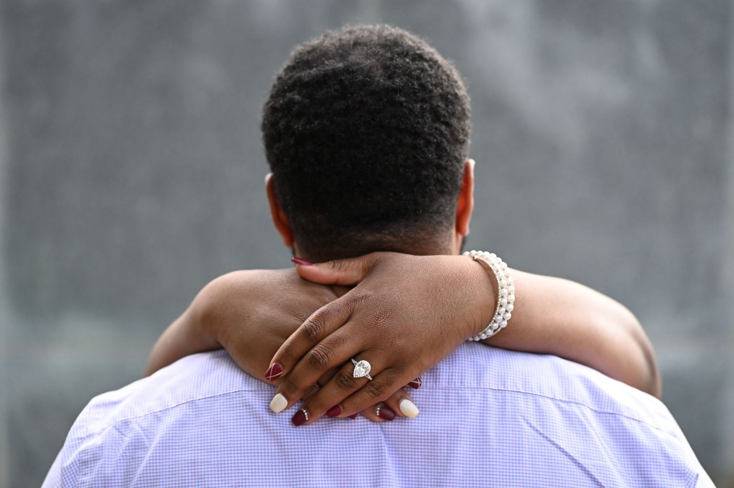 “A close-up of an engaged couple embracing, with the woman’s hands wrapped around her fiancé’s shoulders, showcasing her engagement ring. Ro