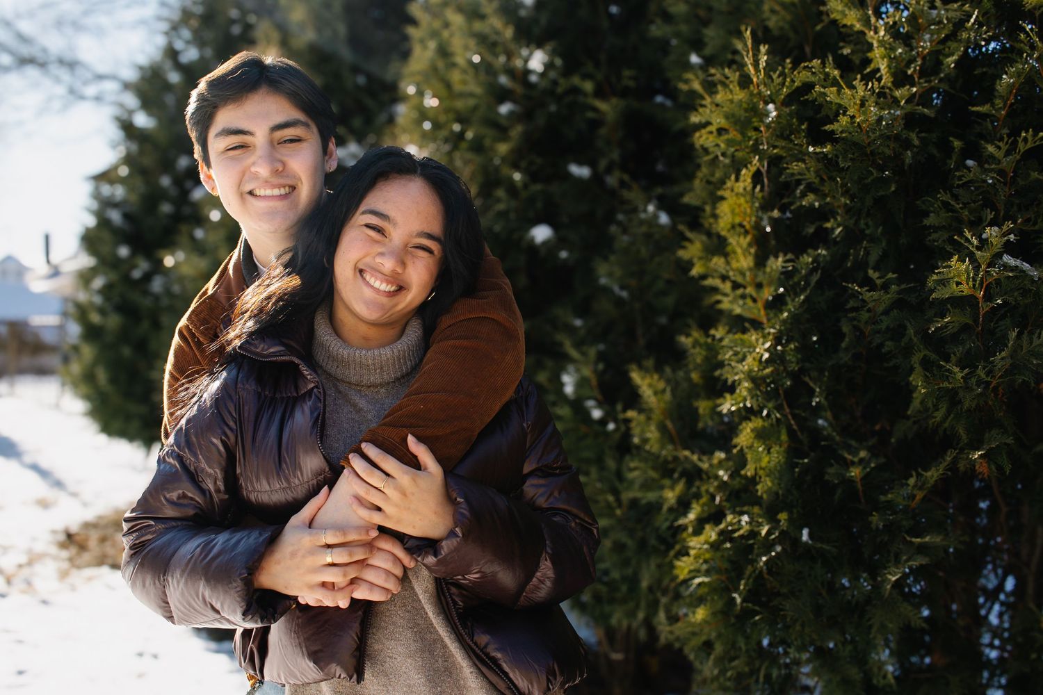 A loving couple shares laughter while embracing in a snowy winter setting next to evergreen trees.