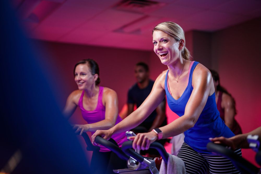 People enjoying an energetic indoor cycling class with vibrant pink and blue lighting.