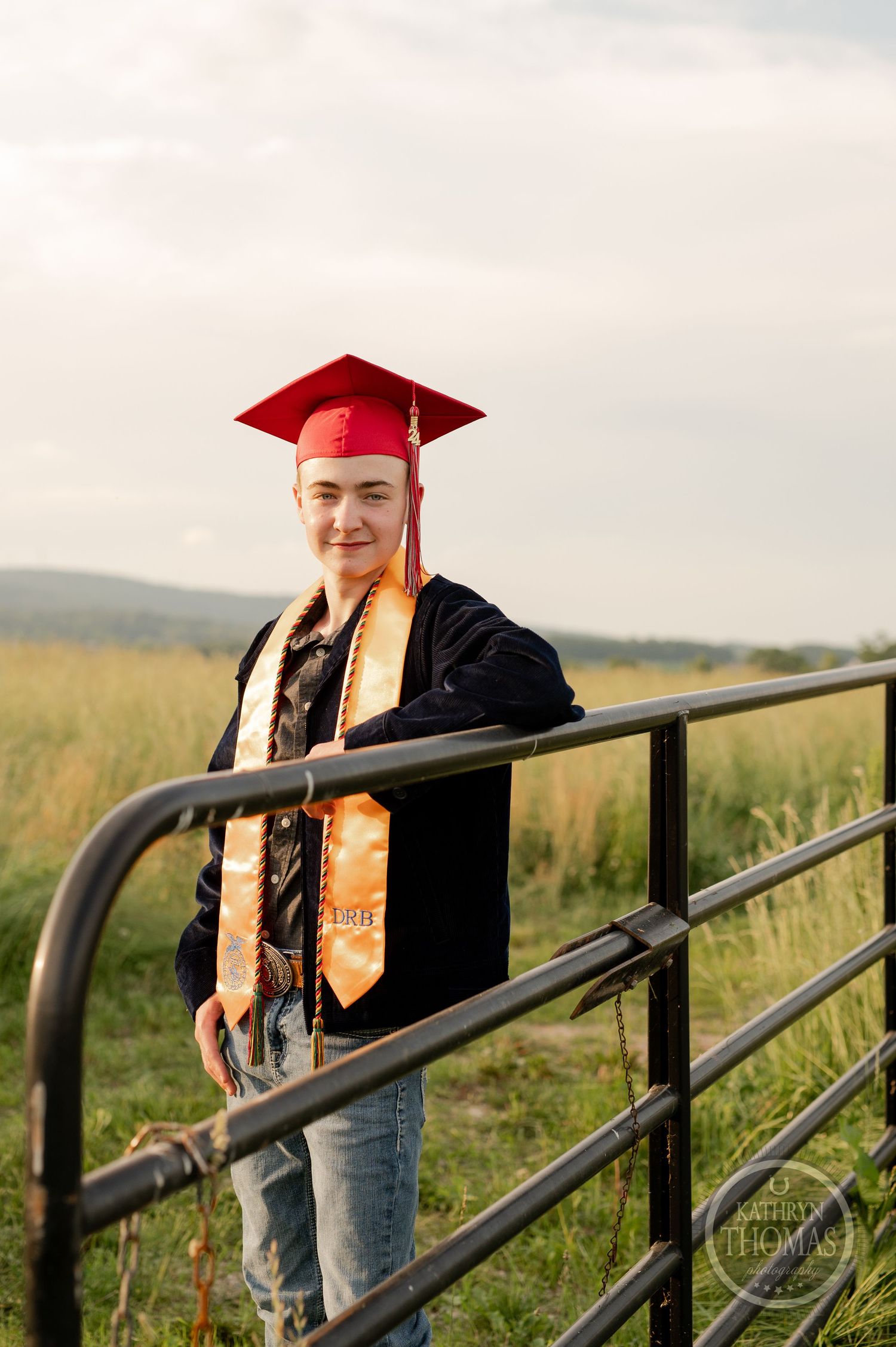 Graduate poses by metal gate in field wearing red cap and gold graduation stole.