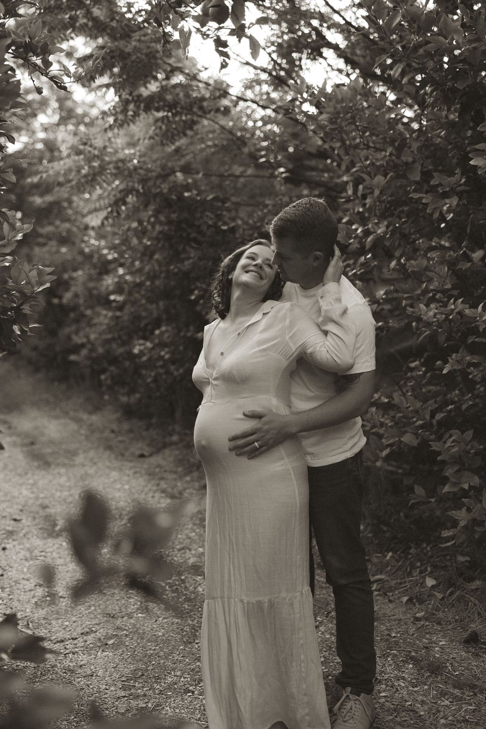 A couple embraces tenderly in a romantic outdoor setting surrounded by lush greenery and trees.