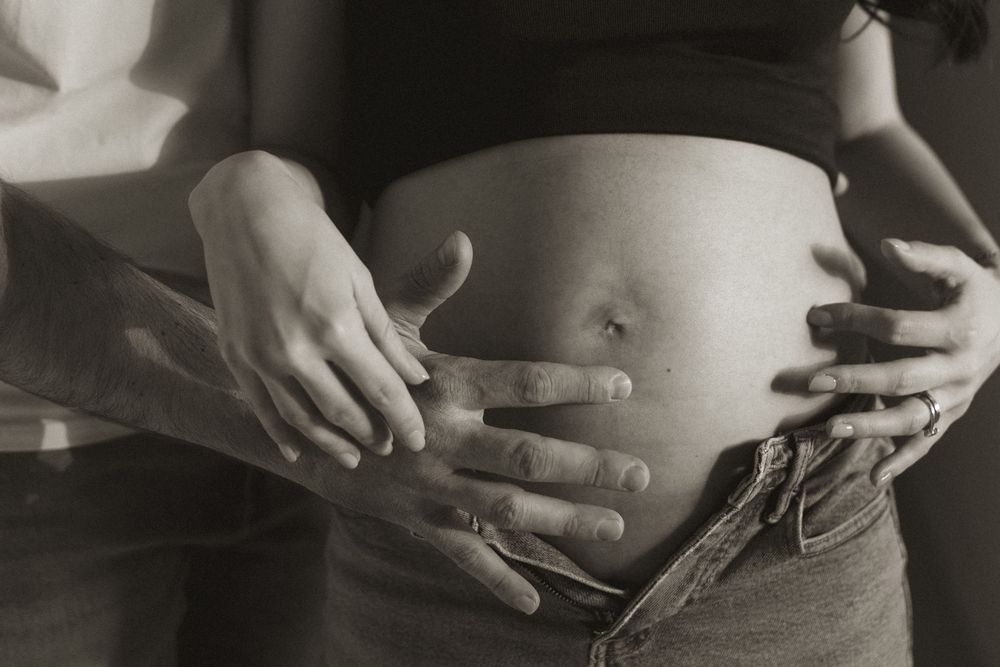 Black and white photo of a pregnant woman's bare baby bump with two pairs of hands gently cradling her belly.