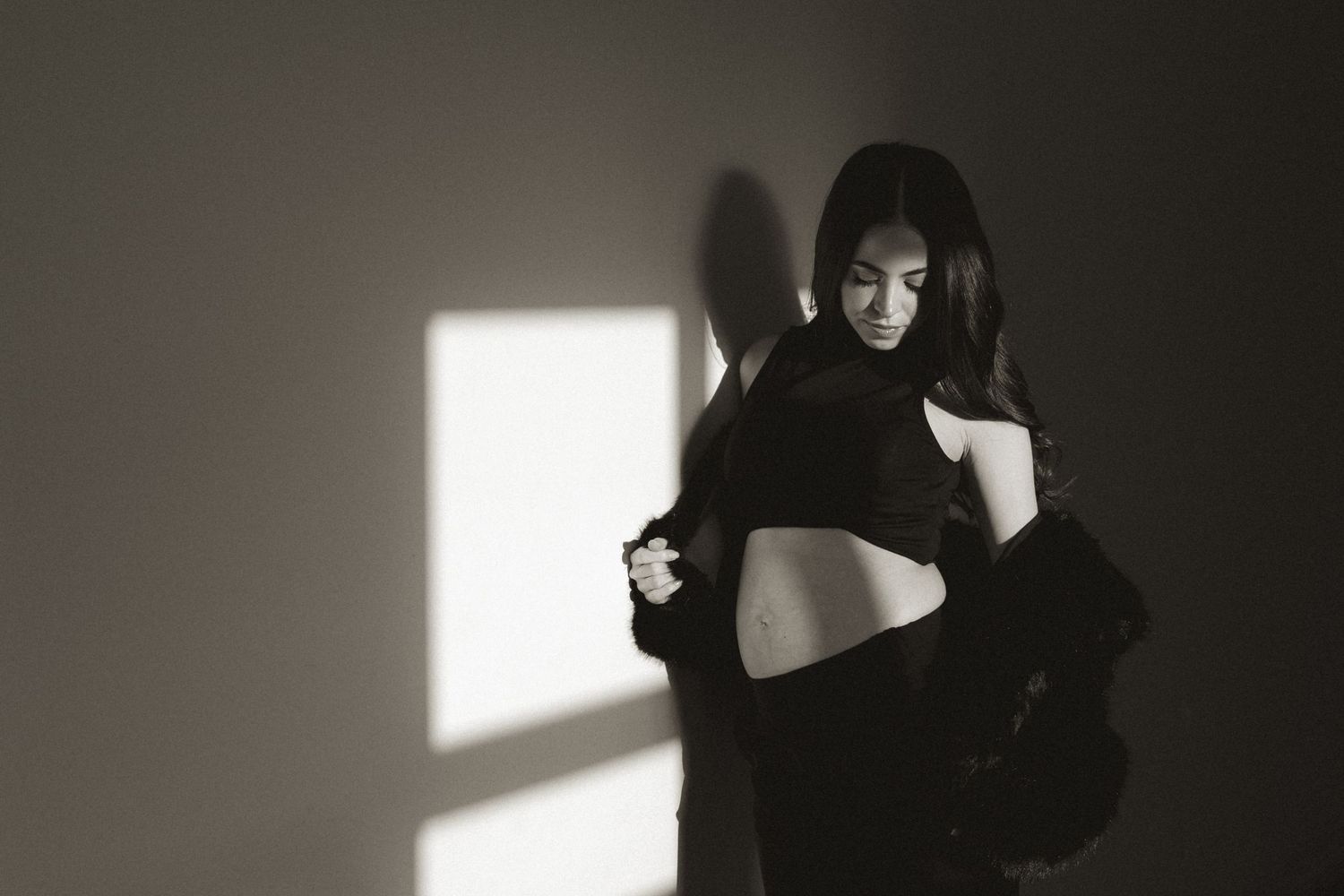 Black and white photo of a woman posing in dramatic light, wearing a cropped top and dark jacket indoors.