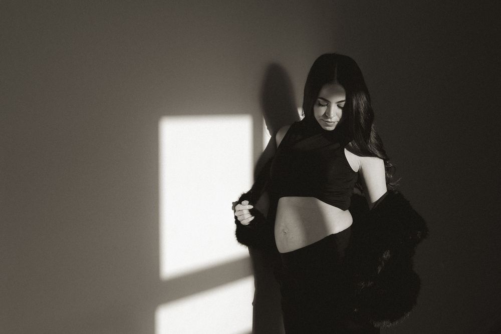 Black and white photo of a woman posing in dramatic light, wearing a cropped top and dark jacket indoors.