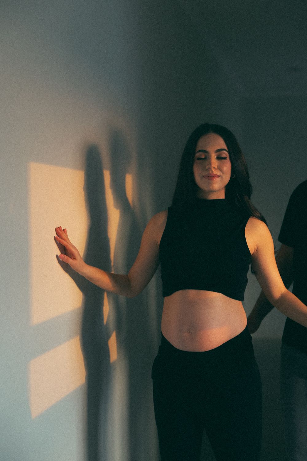 Pregnant woman in black crop top and pants smiling against a wall with warm golden light shadows.