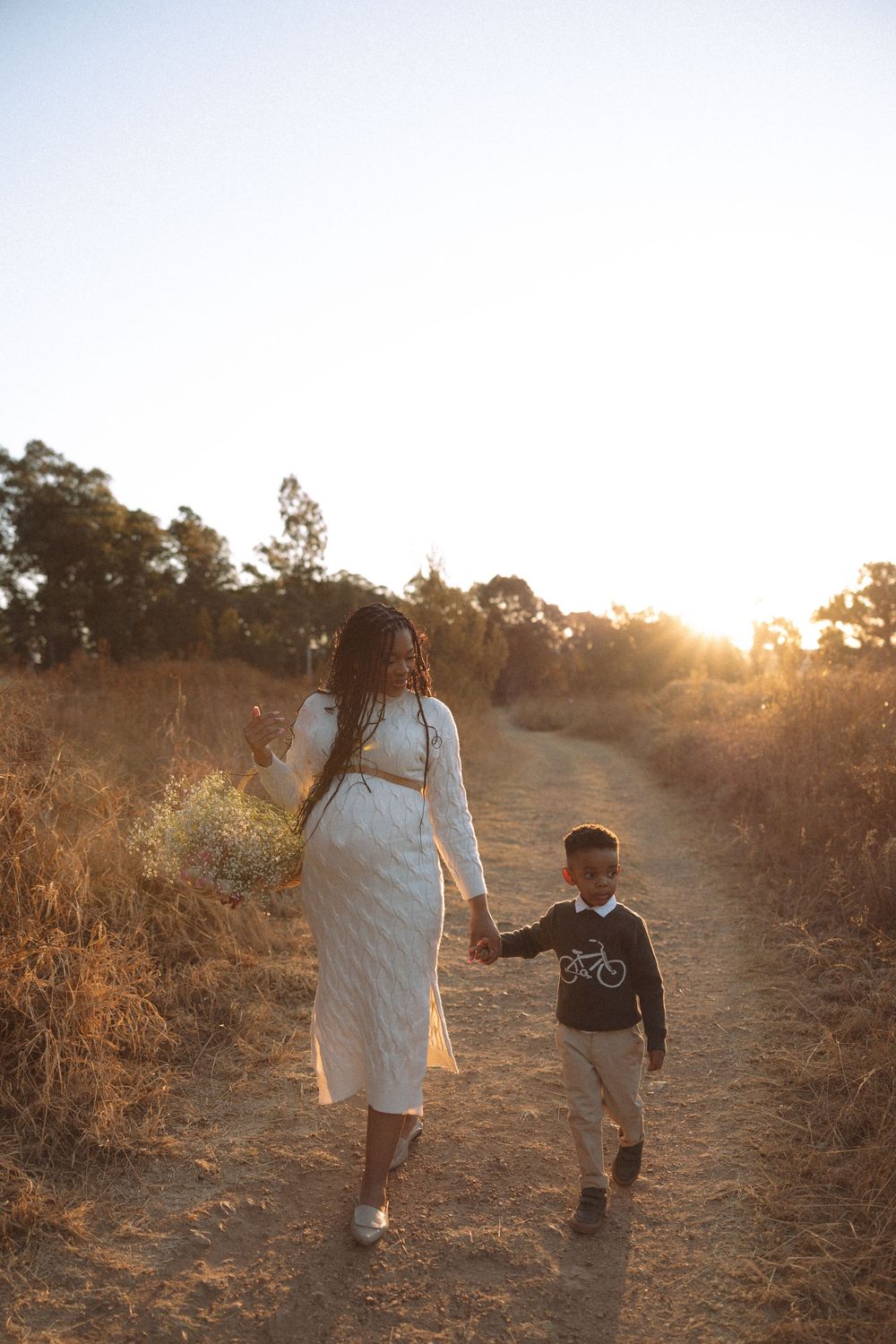 Two people walk hand in hand along a dusty trail during golden hour as the sun sets behind them in a natural setting.